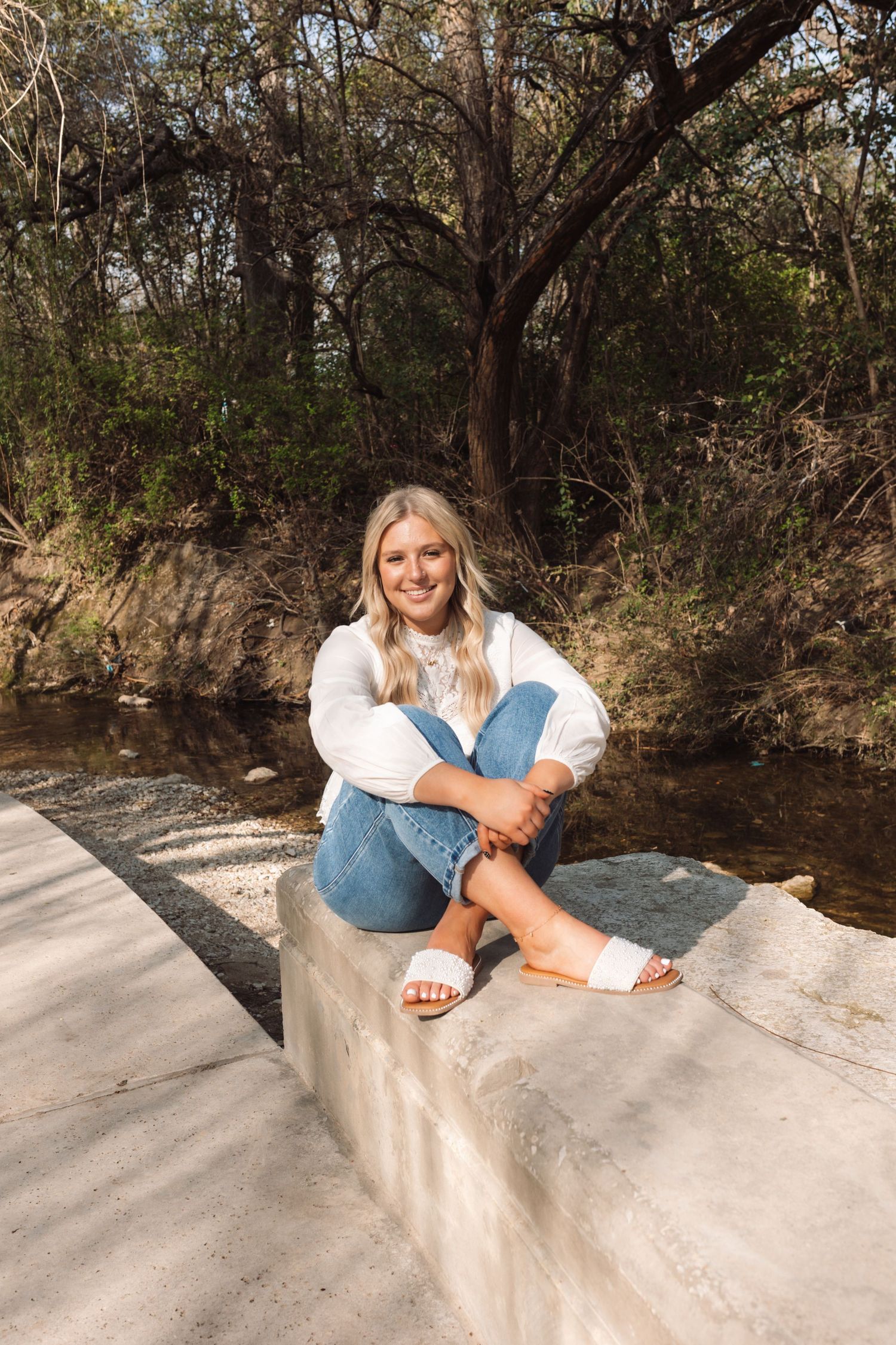 A person in casual attire relaxes on a concrete ledge near a serene creek or river on a sunny day.