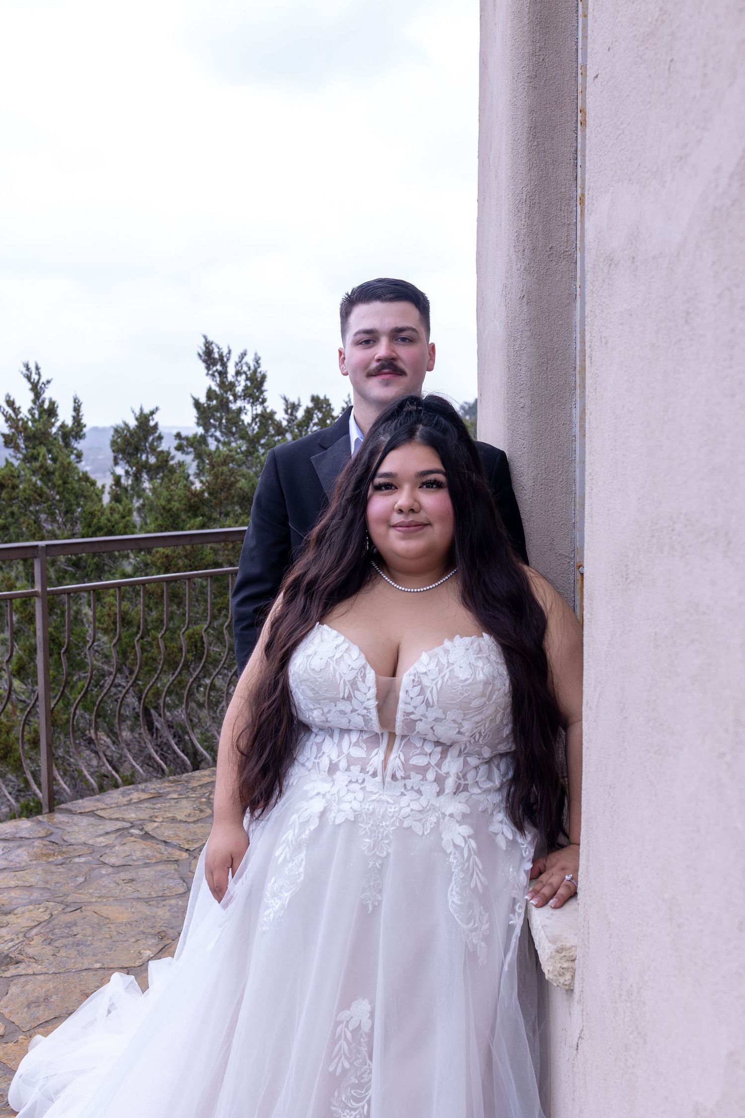 A bride in a strapless white lace wedding gown poses on a balcony with stone columns and greenery in the background.