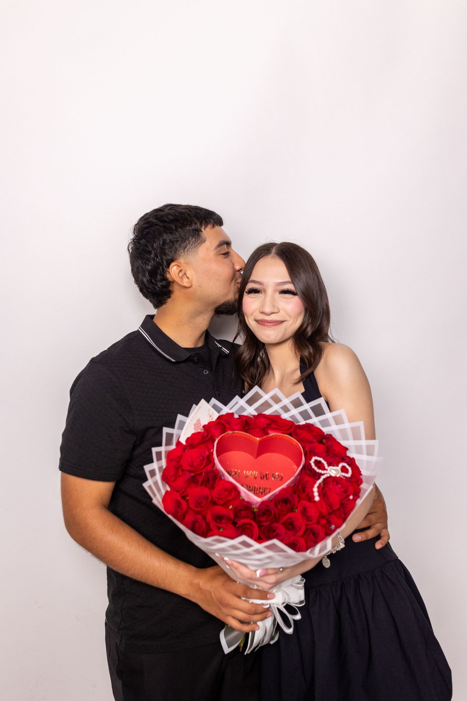 A couple in black outfits poses with a red heart-shaped bouquet of roses against a white backdrop in three romantic shots.