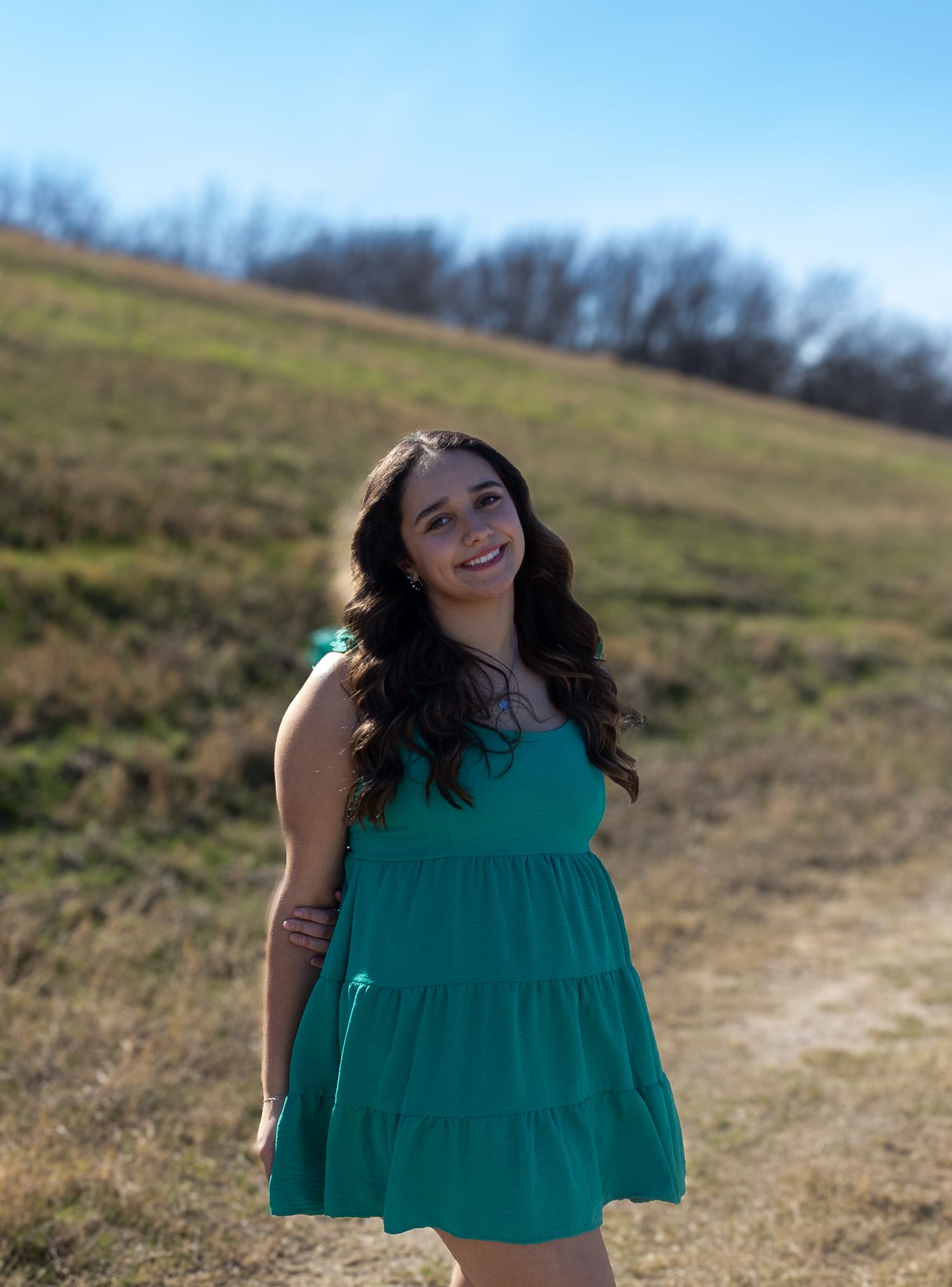 A person in a green sleeveless top poses playfully in a sunny field with blue sky and trees in the background.
