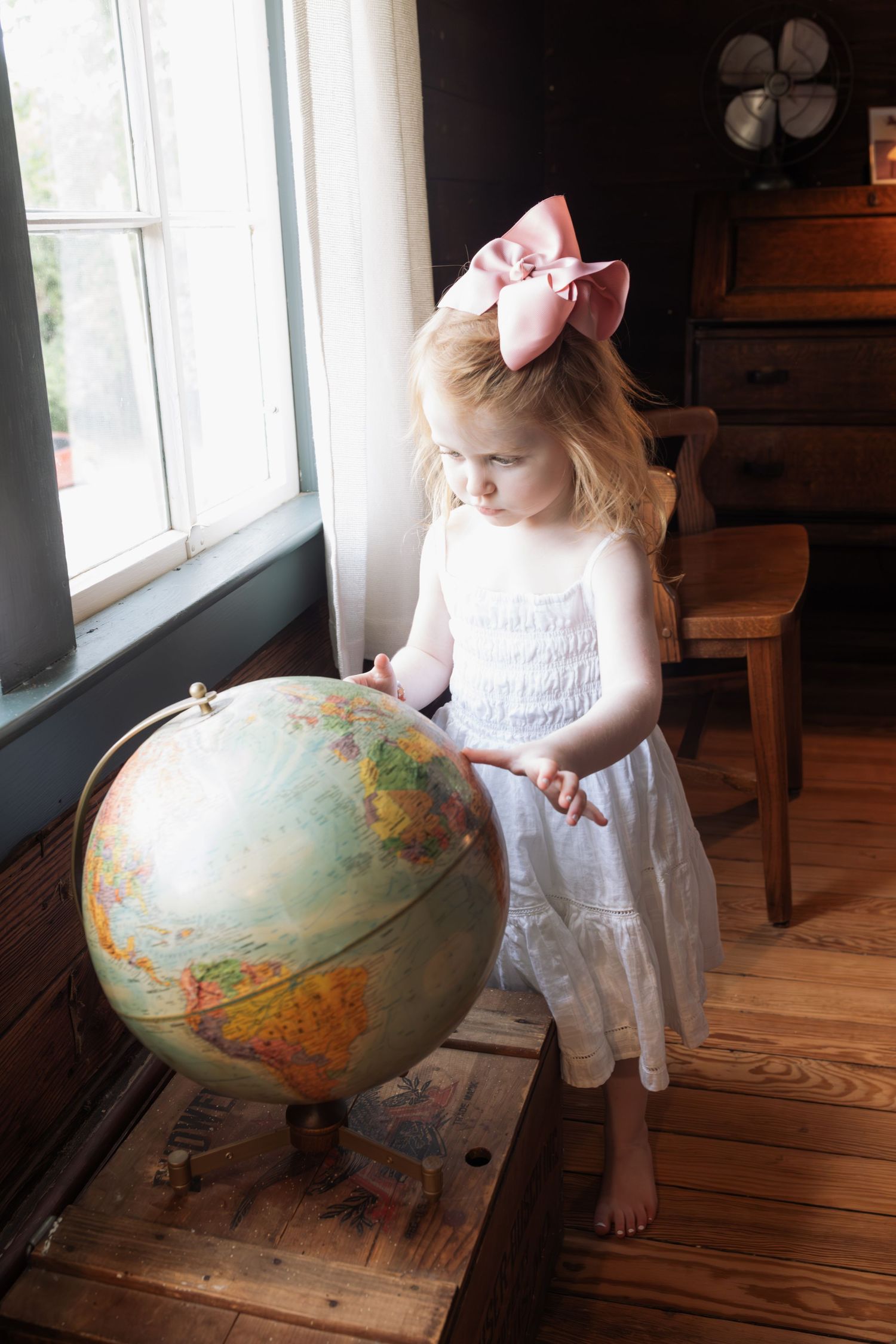 A young girl with a pink bow in her hair points at a globe while standing barefoot in a vintage wooden room.