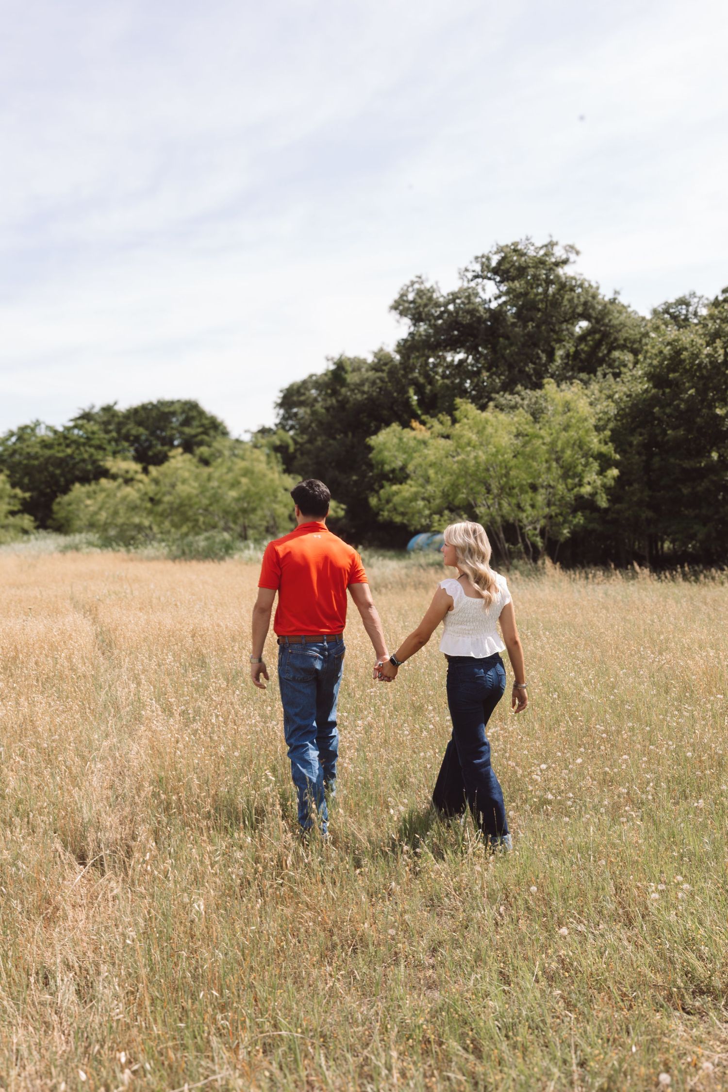 Couple holding hands walking through a golden meadow with trees in the background on a sunny day.