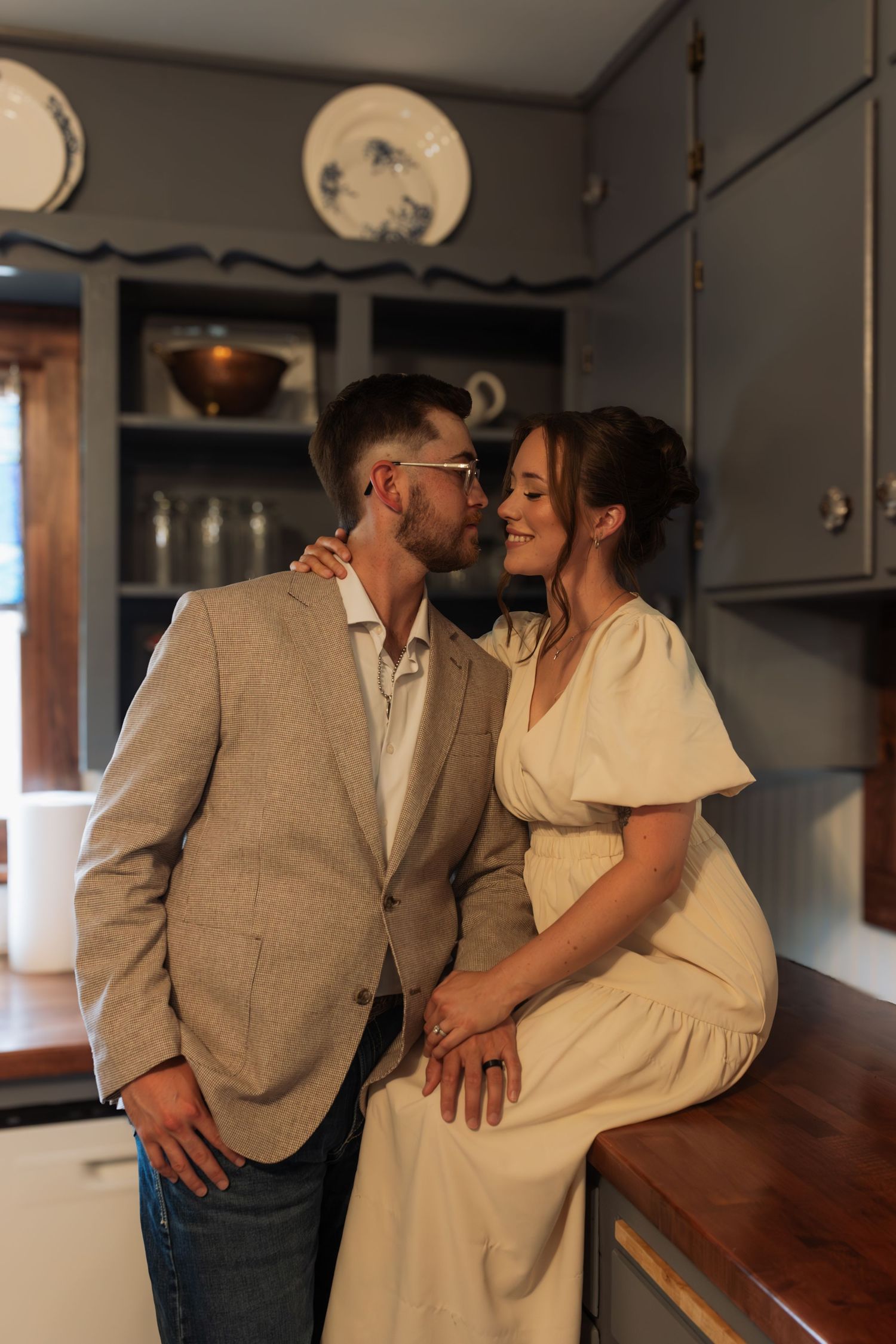 Couple sharing an intimate moment in a vintage kitchen, bride in cream dress seated on counter.