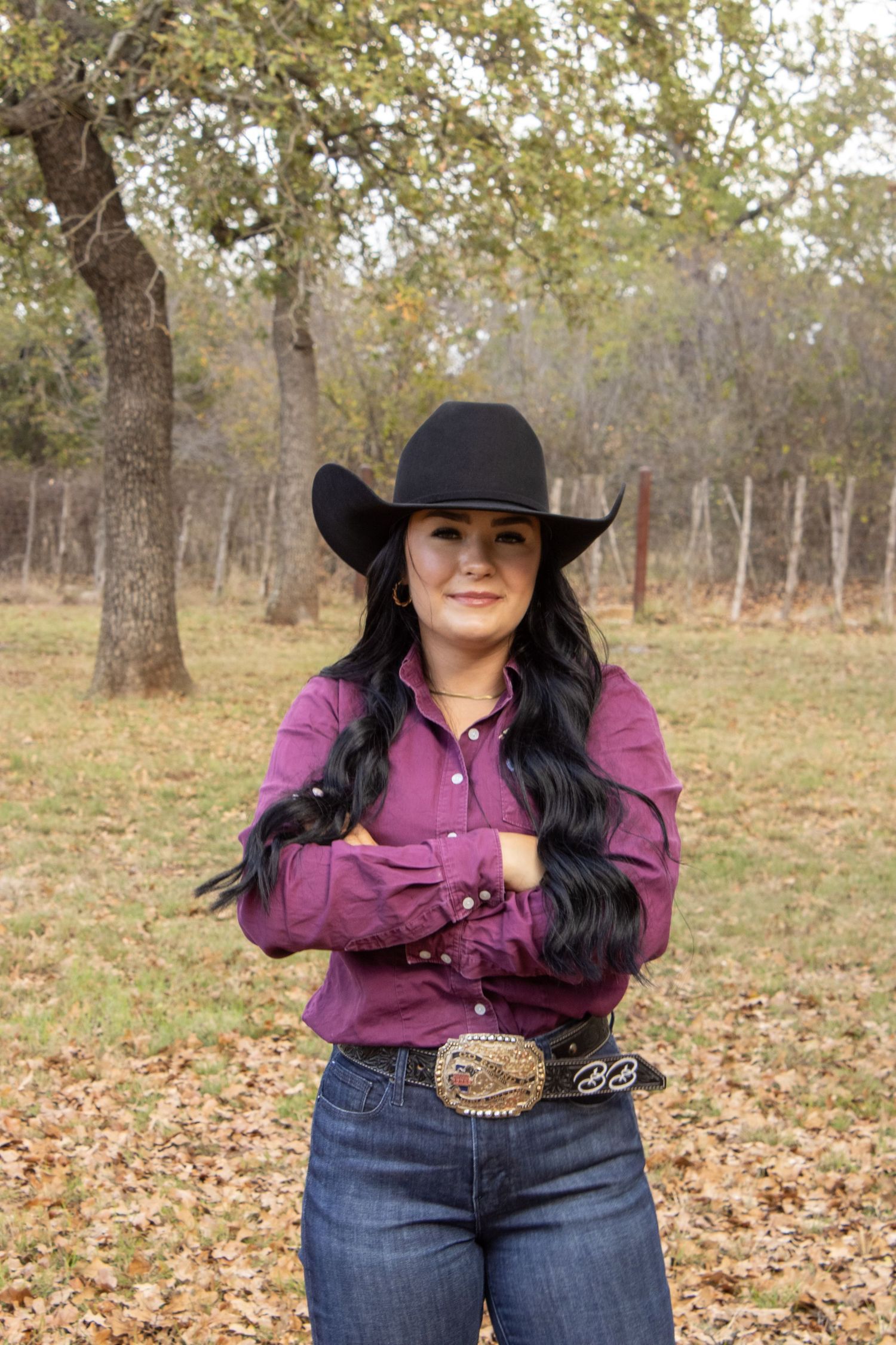 Western attire featuring purple shirt, ornate belt buckle and black cowboy hat against fall foliage background.