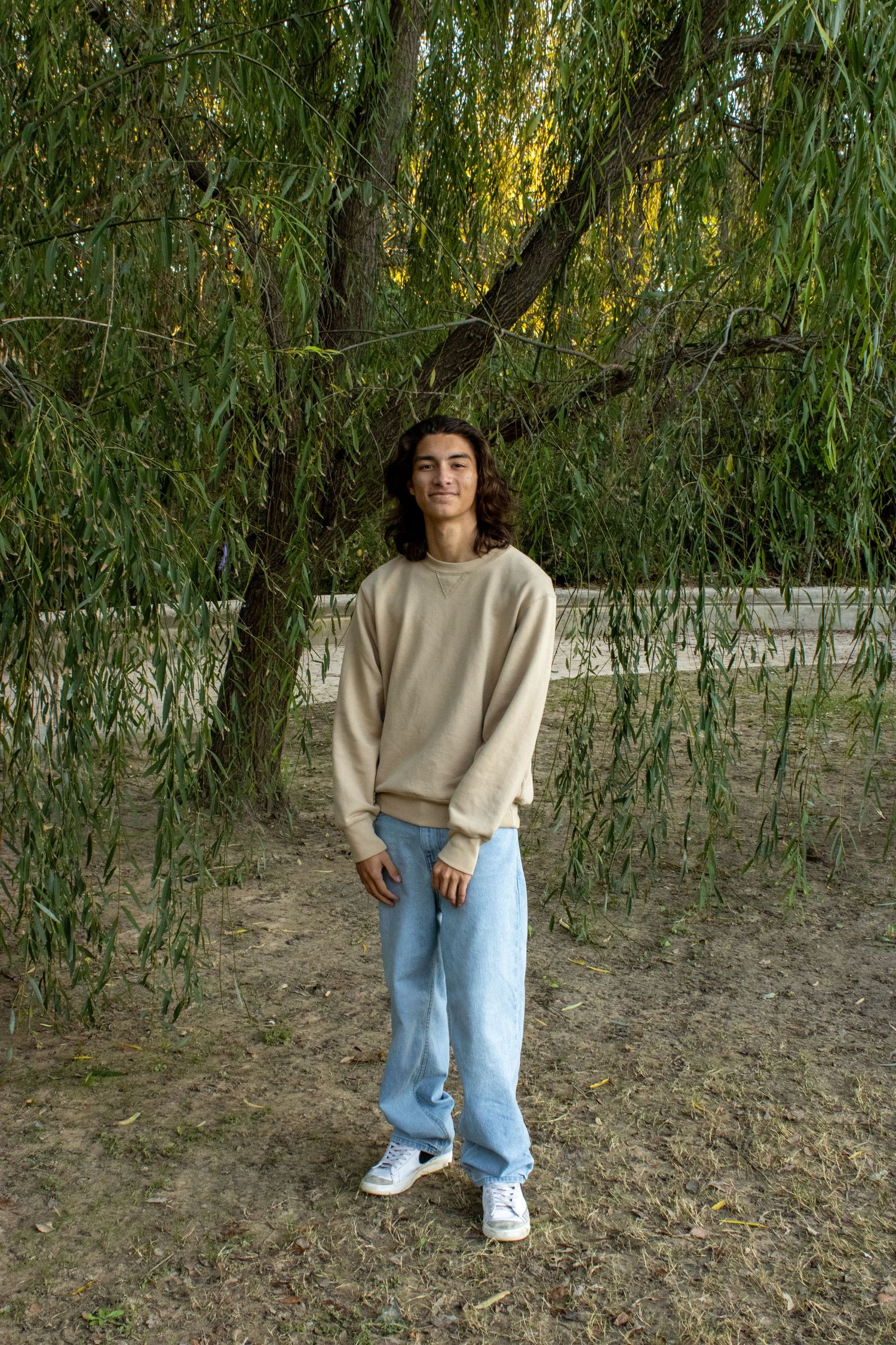 A person in a beige sweater and light blue jeans stands beneath drooping willow tree branches in a natural setting.