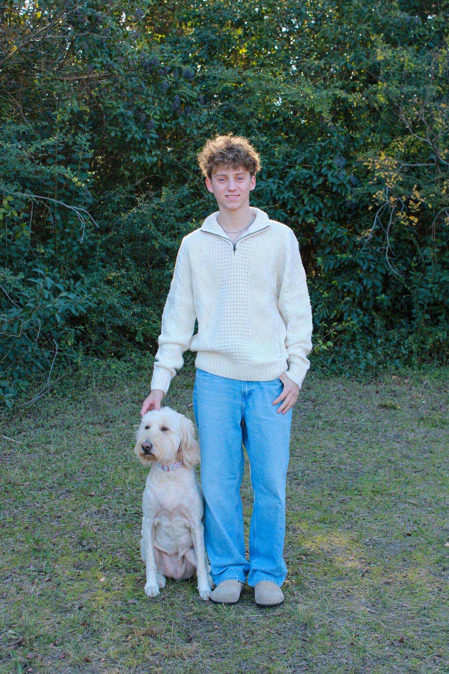 A person in a white sweater and blue jeans stands with a fluffy cream-colored dog in a grassy outdoor setting.