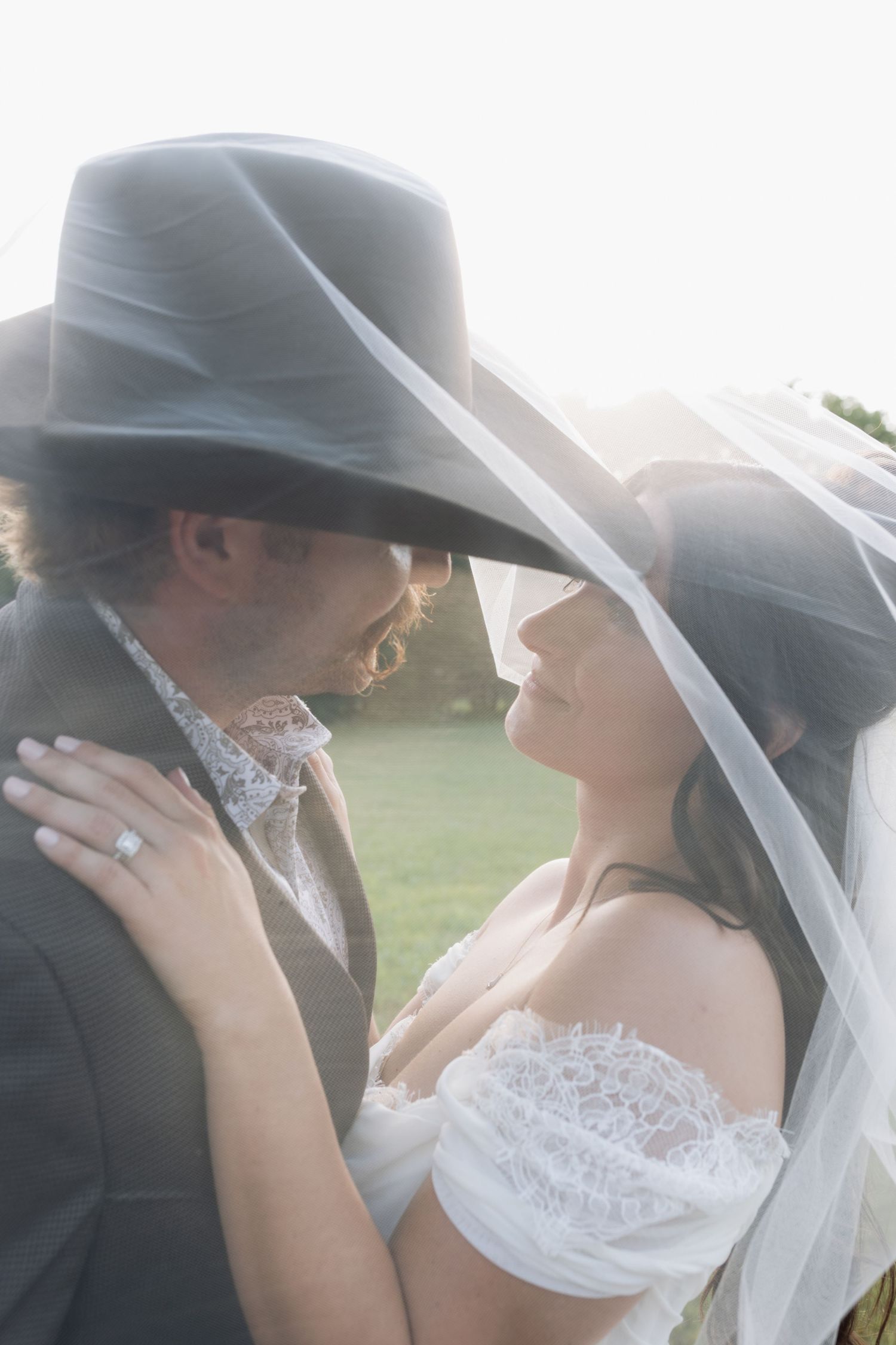 Romantic close-up of bride and groom nearly kissing under a wide-brim cowboy hat and flowing wedding veil outdoors.
