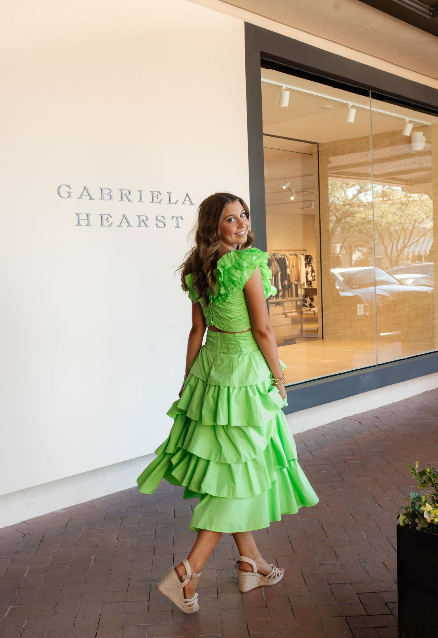Woman in bright green ruffled midi dress and wedge sandals posing outside Gabriela Hearst boutique.