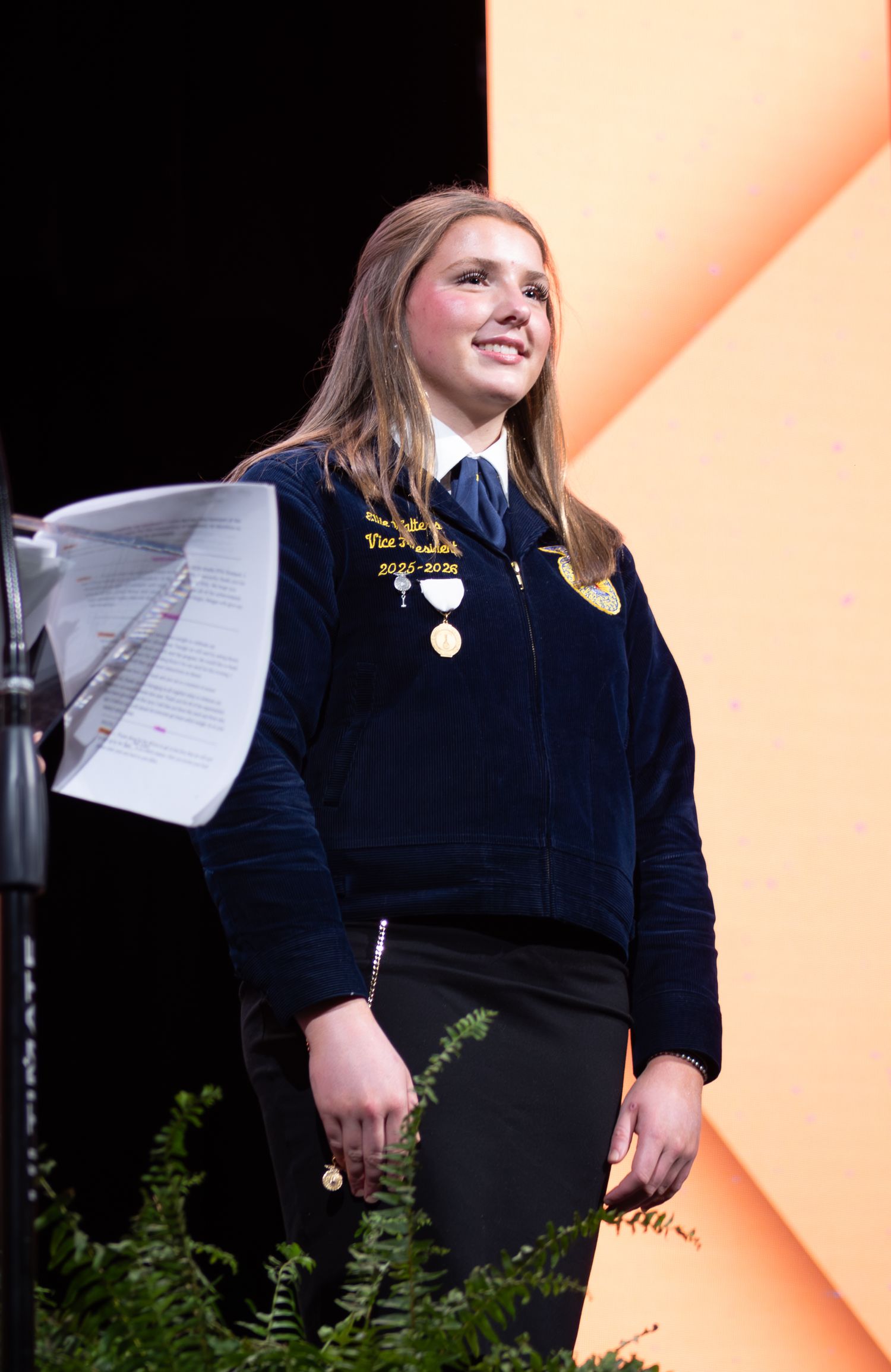 Young woman in navy FFA jacket with medals smiles on stage during an FFA convention ceremony.