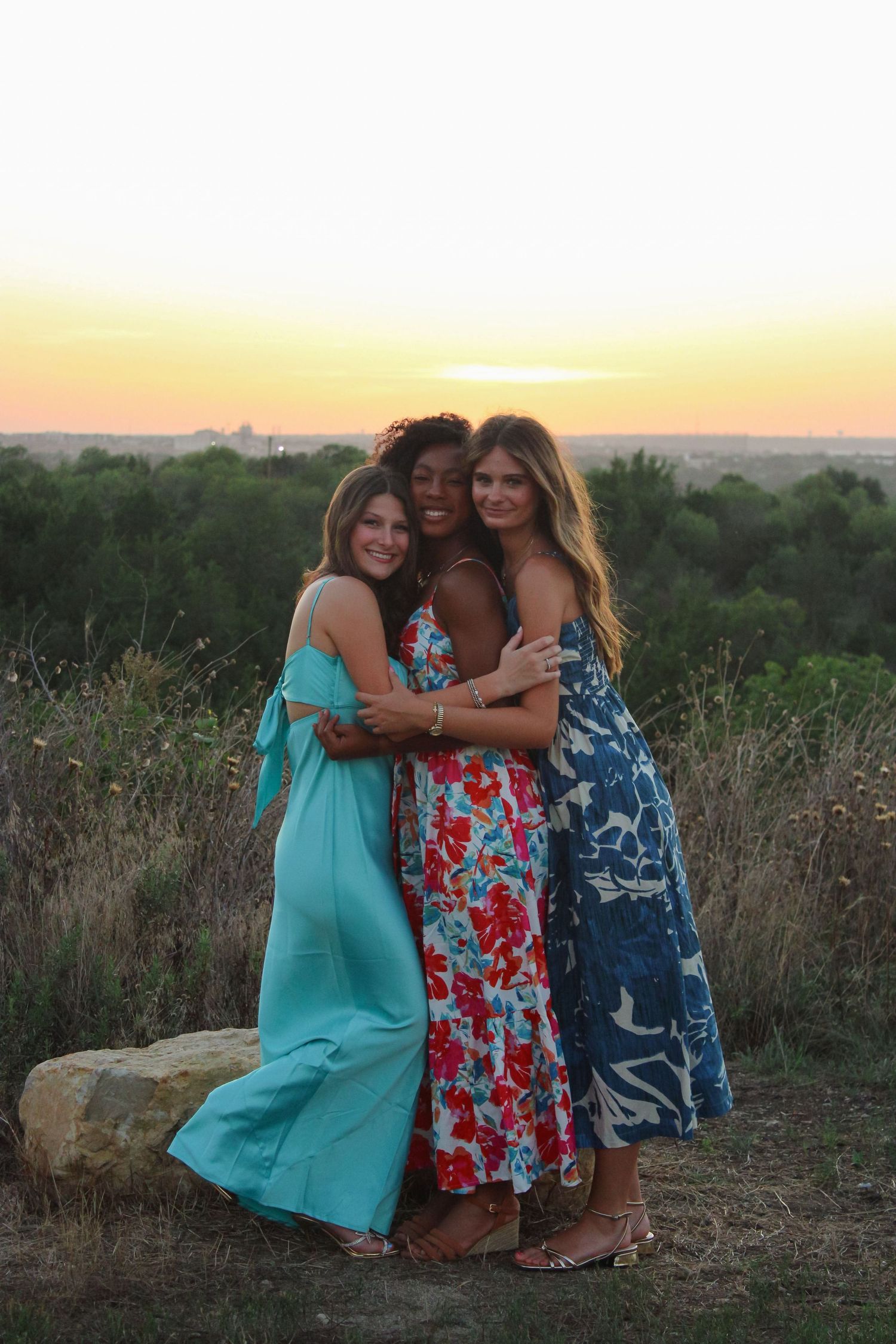 Group embracing while wearing flowing dresses in a sunset field.