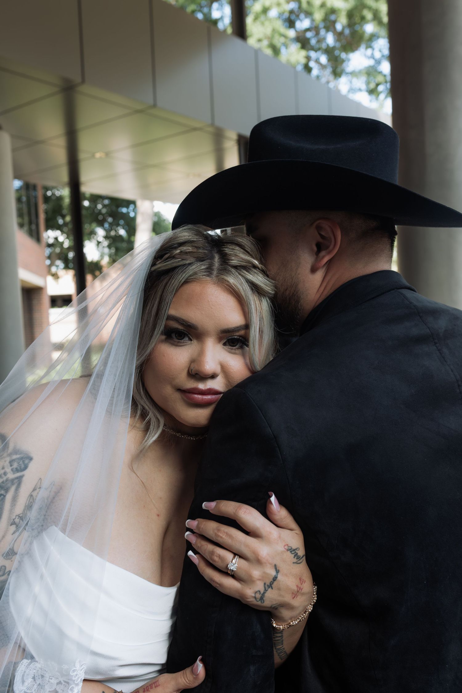 Bride in white dress and veil embraces groom in black suit and cowboy hat outdoors at wedding ceremony.