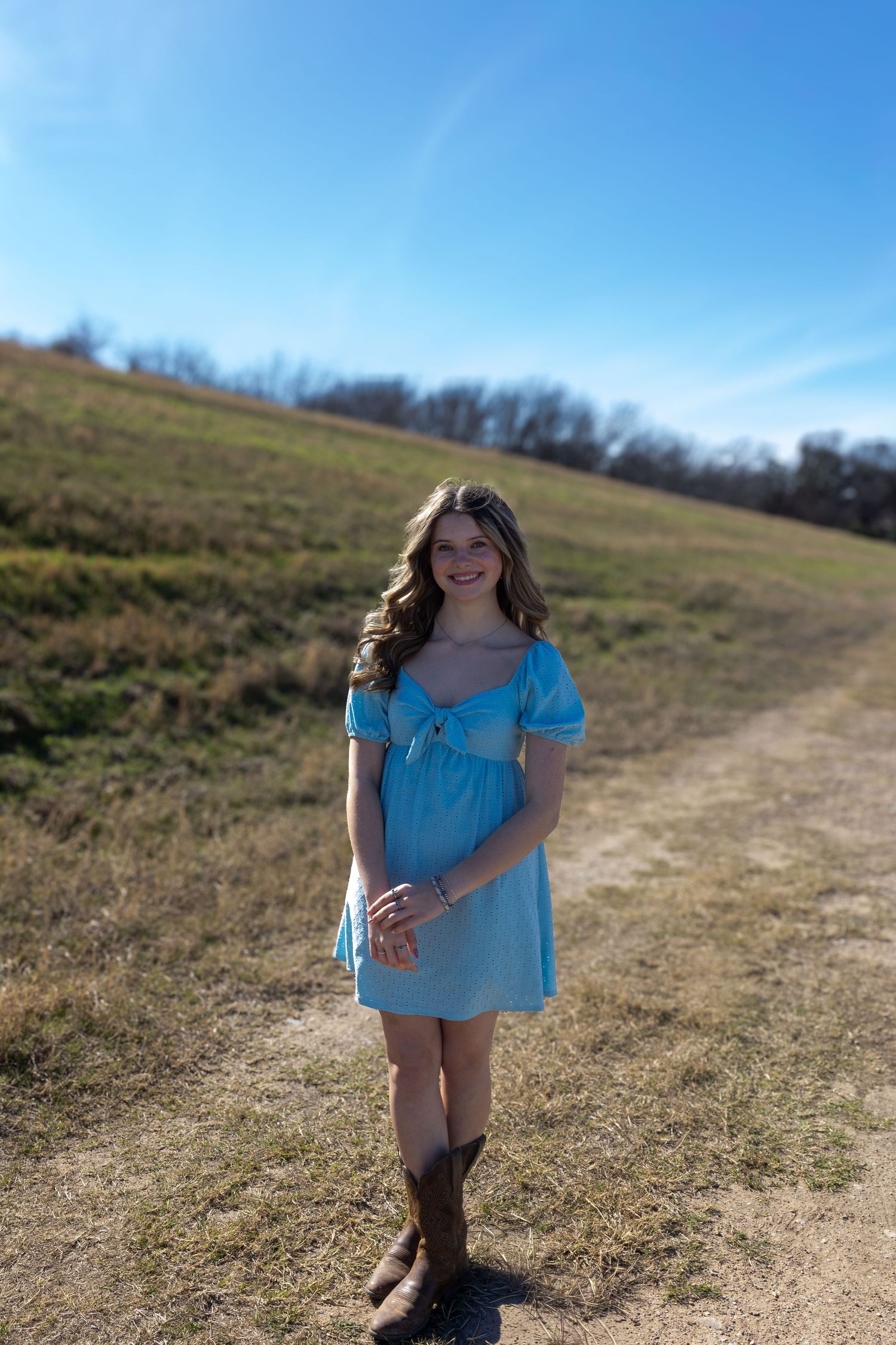 A person in a light blue dress walks along a dirt path on a sunny hillside during a bright afternoon.