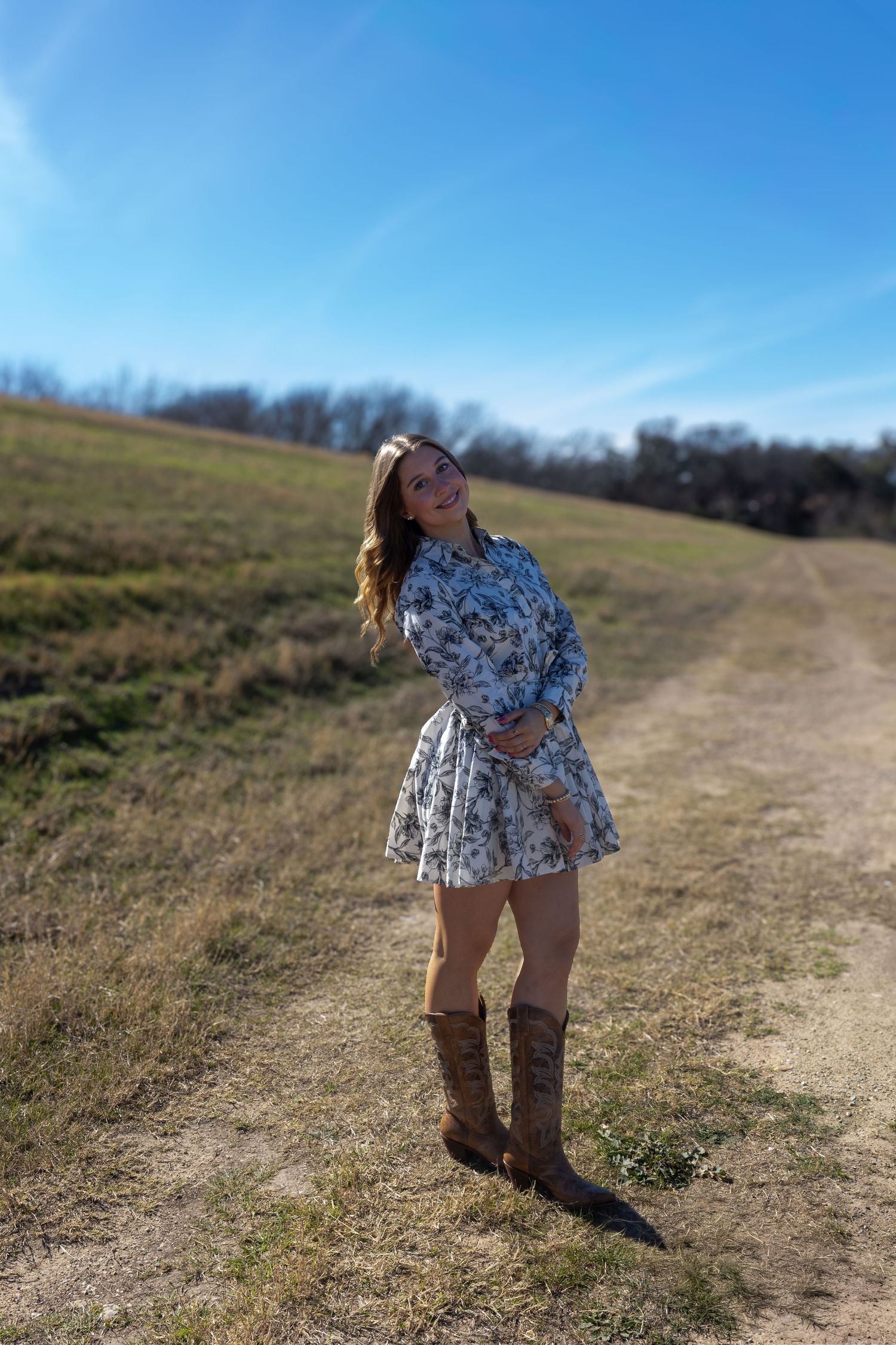 Someone wearing a floral print dress poses on a winding dirt trail through a grassy field on a sunny day.