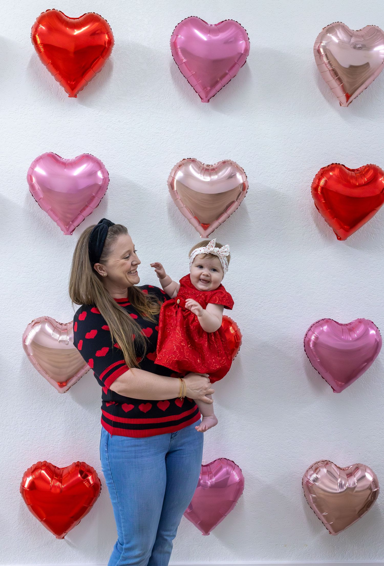 A mother and baby in matching red heart sweaters pose against a wall decorated with pink and red heart balloons.