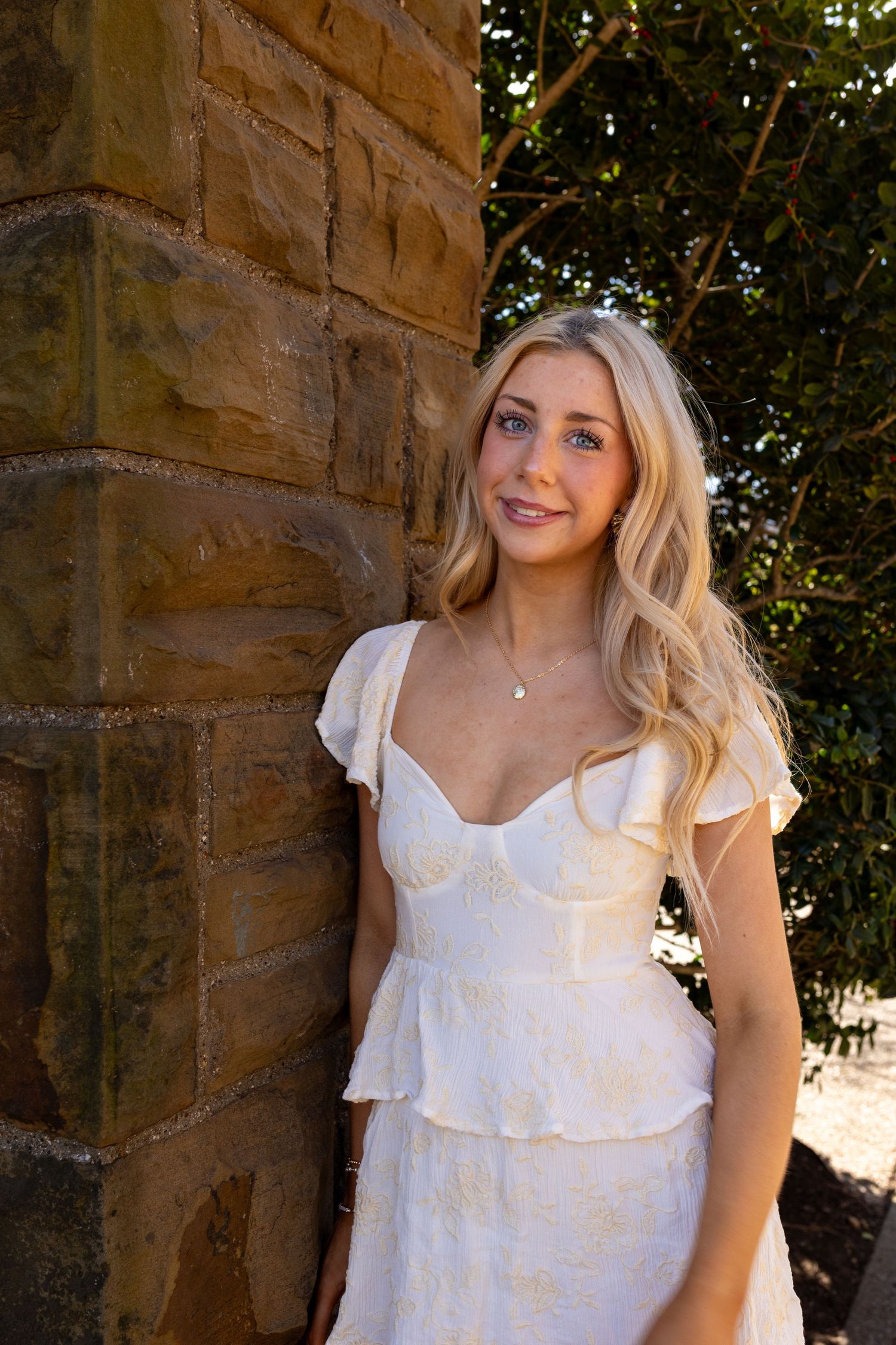 A woman in a white summer dress poses against a sandstone wall in natural outdoor lighting.