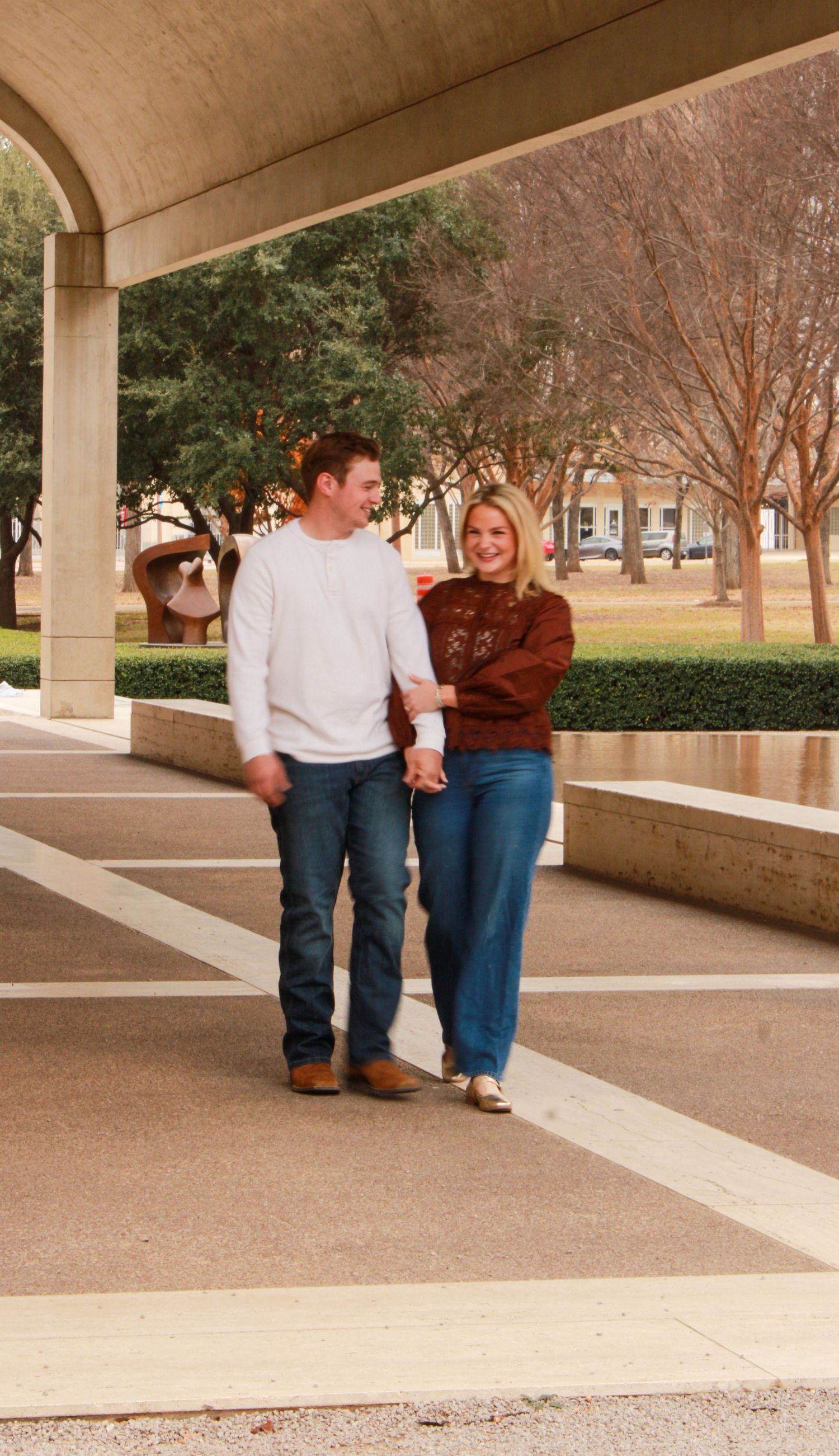 A couple in casual sweaters and jeans walks together under a covered walkway on a college campus.