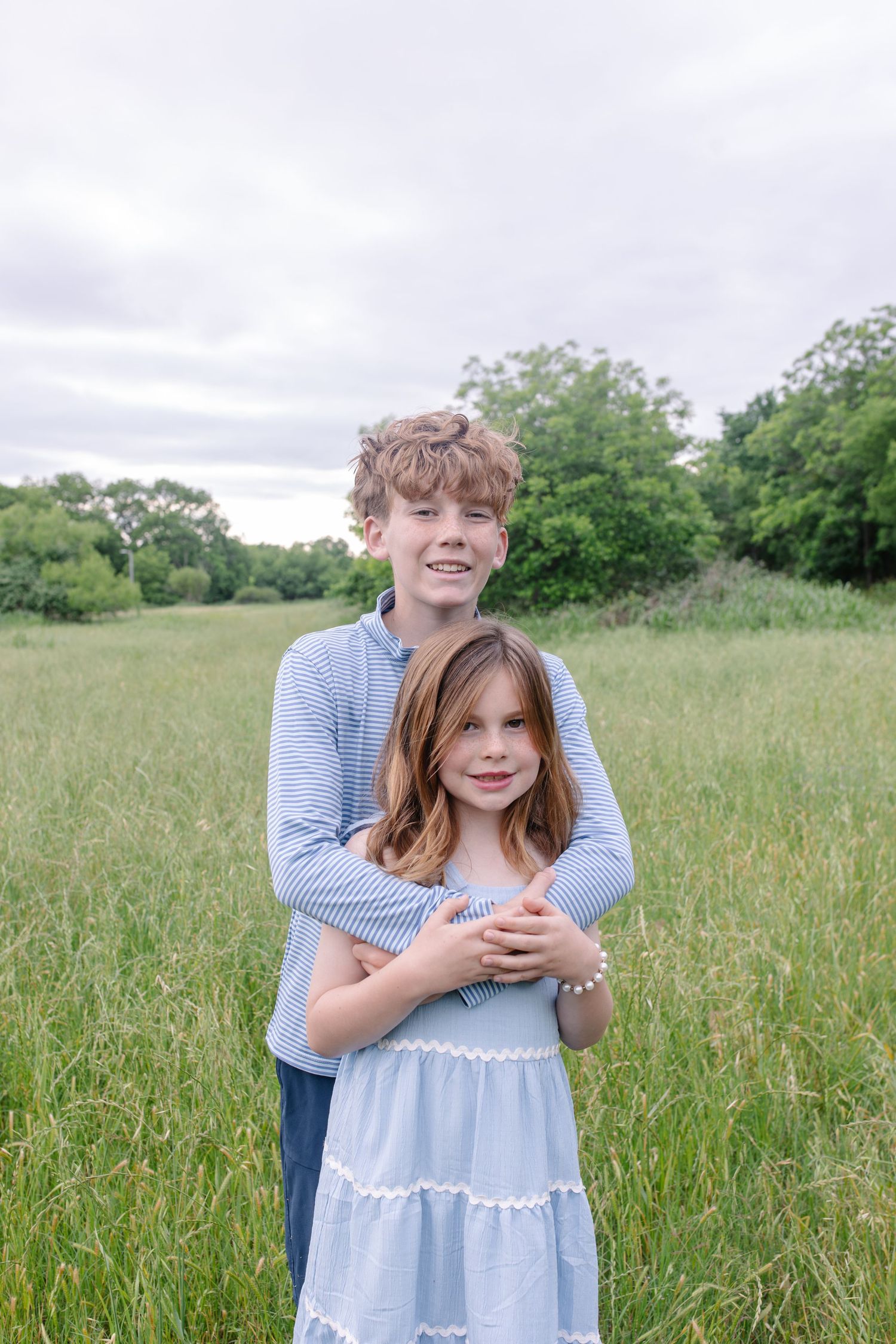 A smiling teenage boy hugs a young girl in a blue dress while standing in a lush green field.