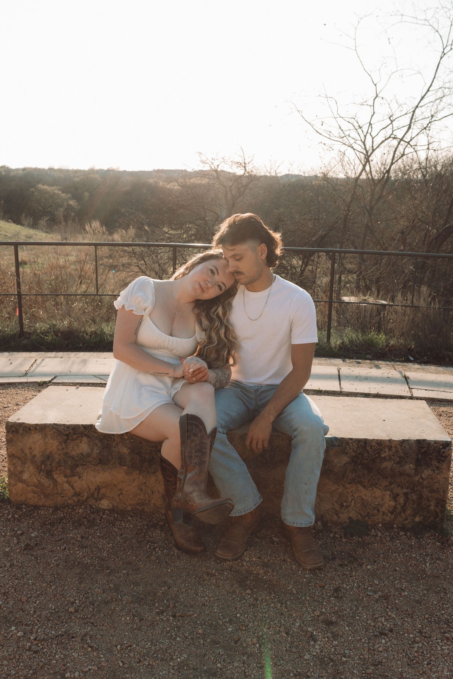 A couple in casual attire sits together on a stone wall at sunset sharing an intimate moment in nature.