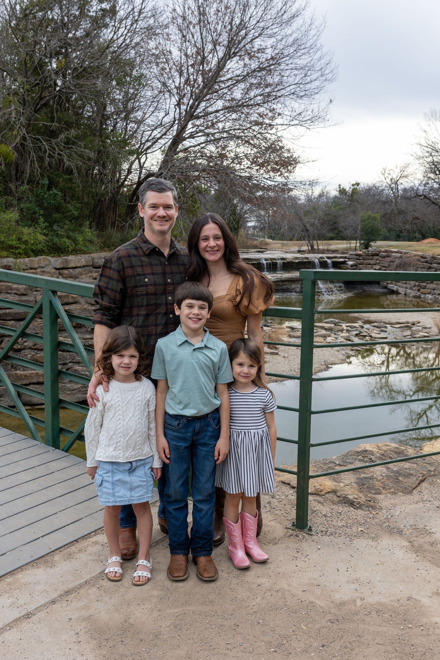 Family standing together on wooden bridge over pond in park on cloudy day.