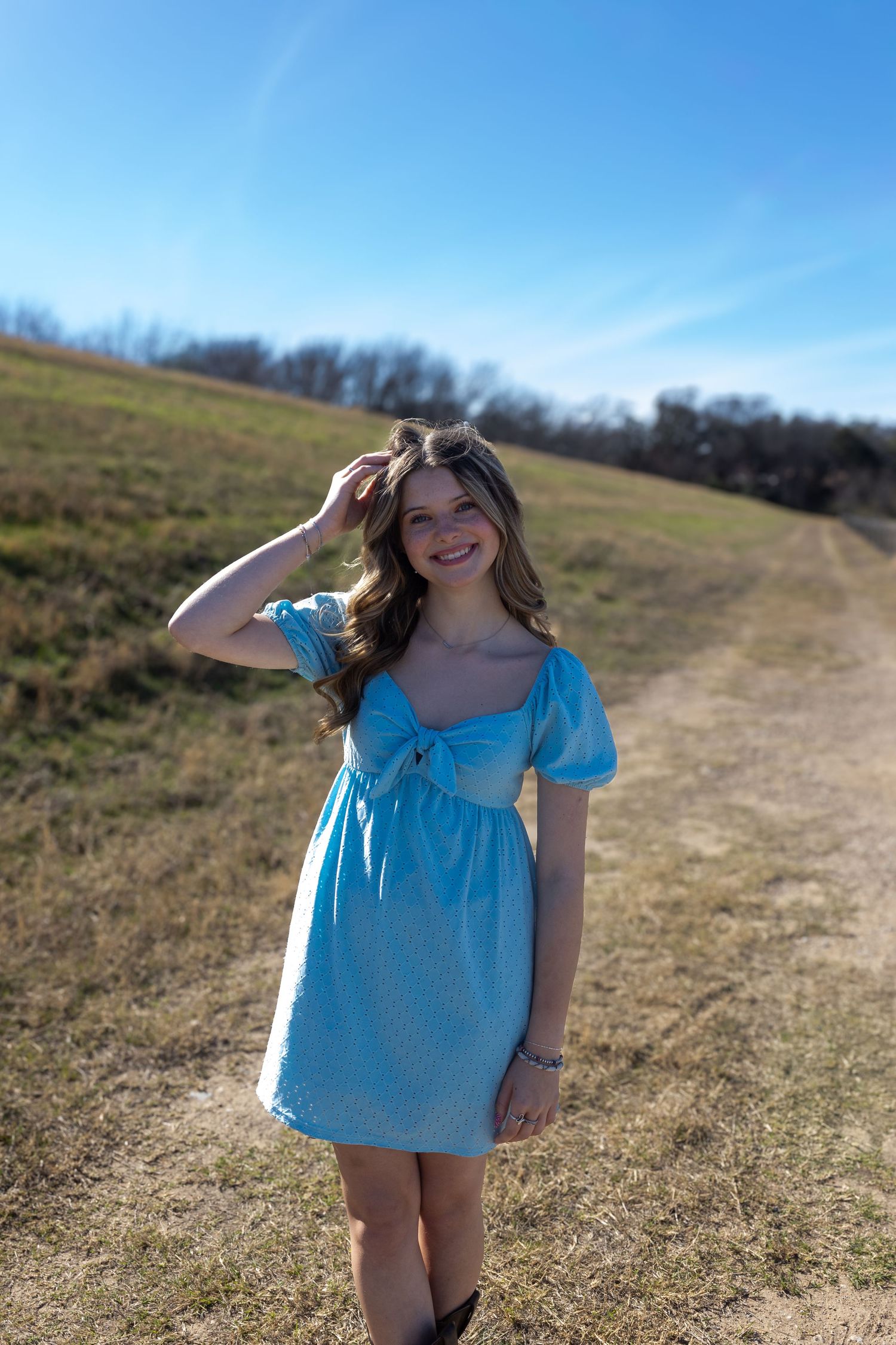 A person in a light blue dress stands on a grassy hillside path against a bright blue sky on a sunny day.