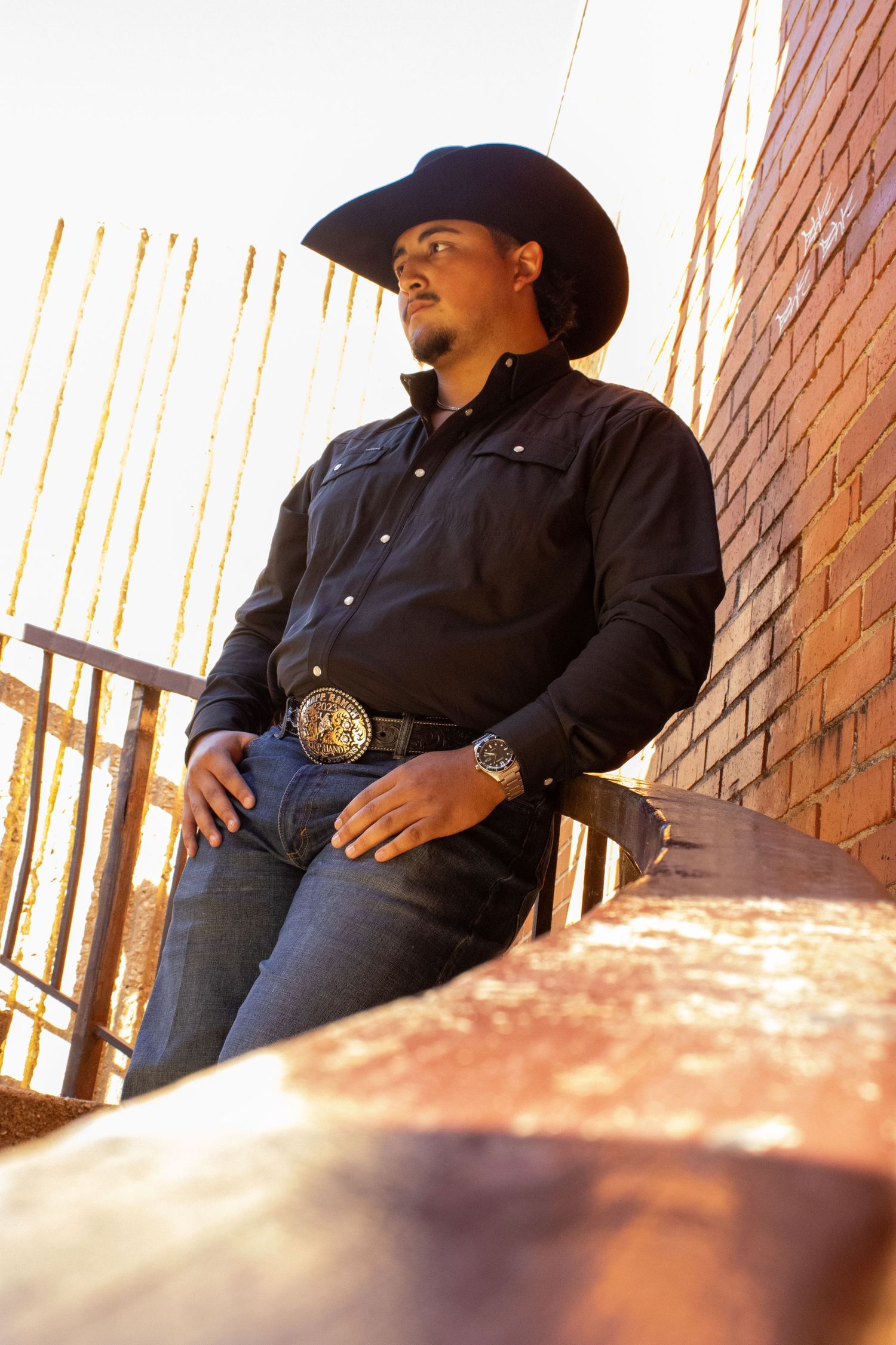 A person in western attire sits against a brick wall in warm sunset lighting.
