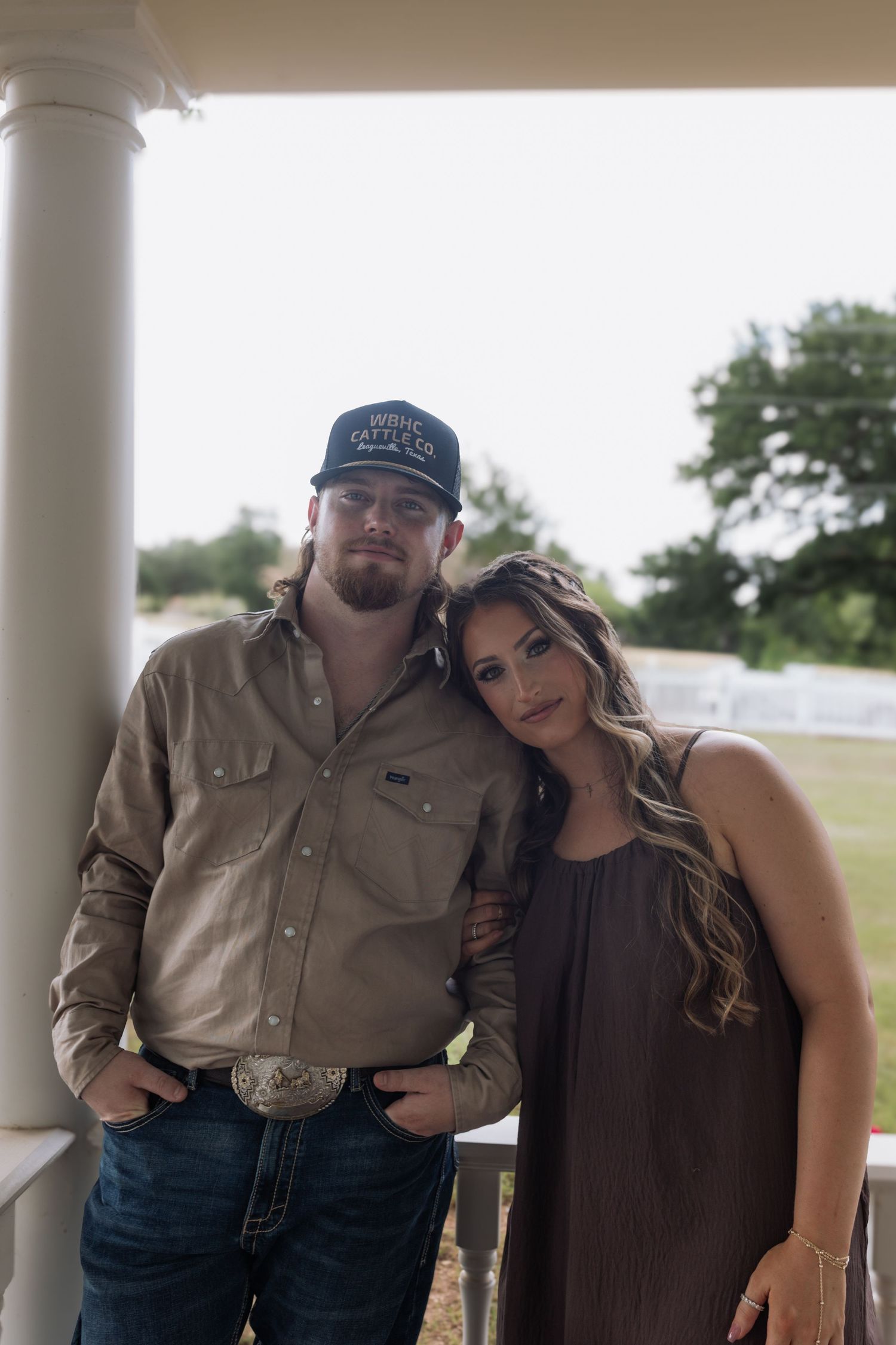 A couple poses on a porch, the man in a cap and jeans, the woman in a brown dress leaning on him.