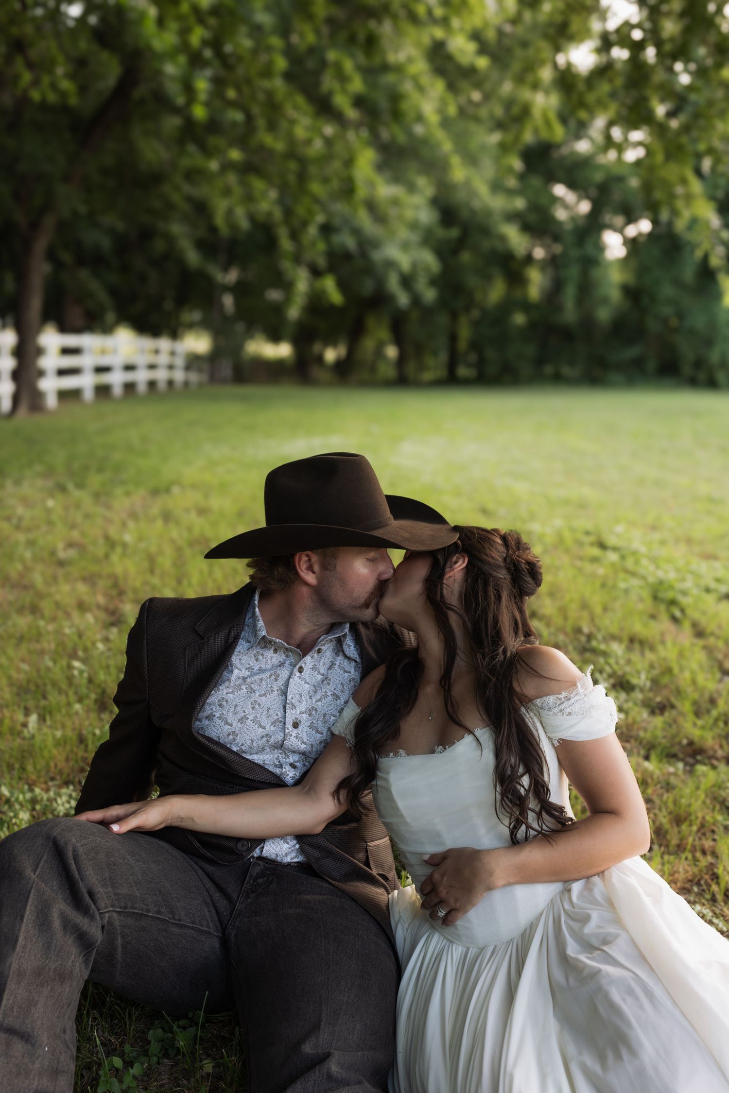 A couple in wedding attire shares a romantic kiss while sitting on grass at a rustic farm with a white fence backdrop.