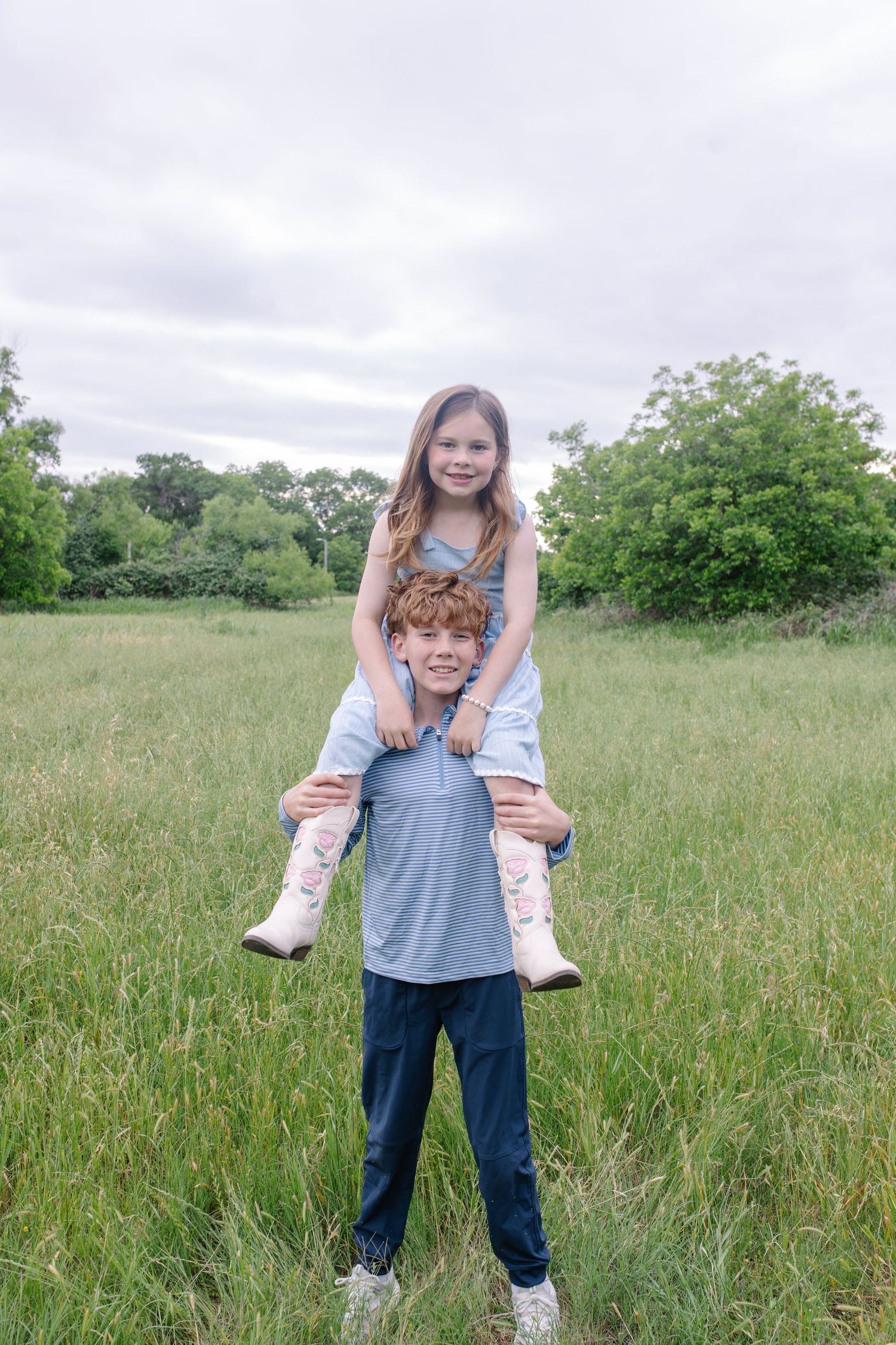 A smiling girl sits on a boy's shoulders in a green meadow, both children laughing outdoors under a cloudy sky.