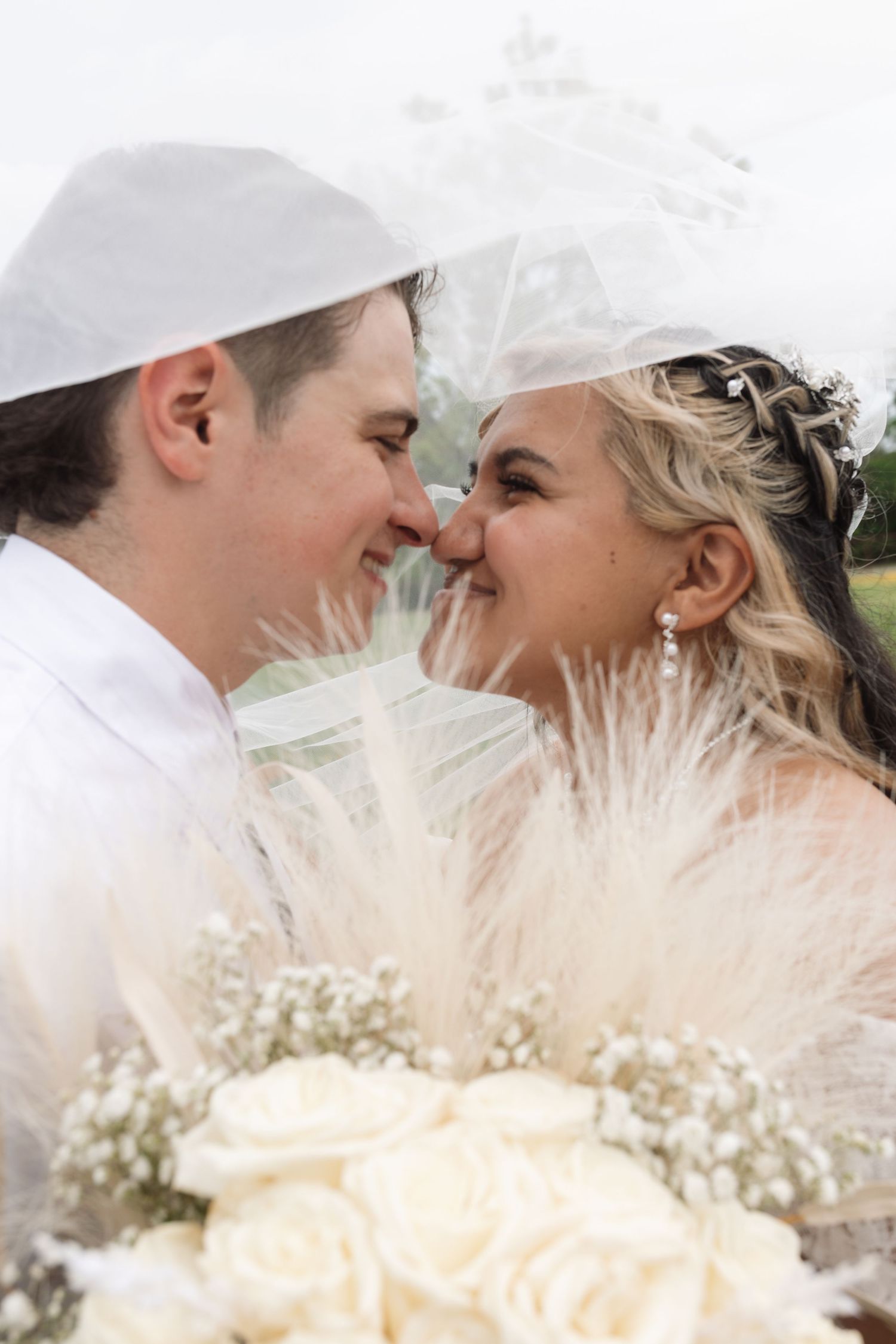 A bride and groom share a romantic nose-to-nose moment under a veil, holding a white rose bouquet at their wedding.