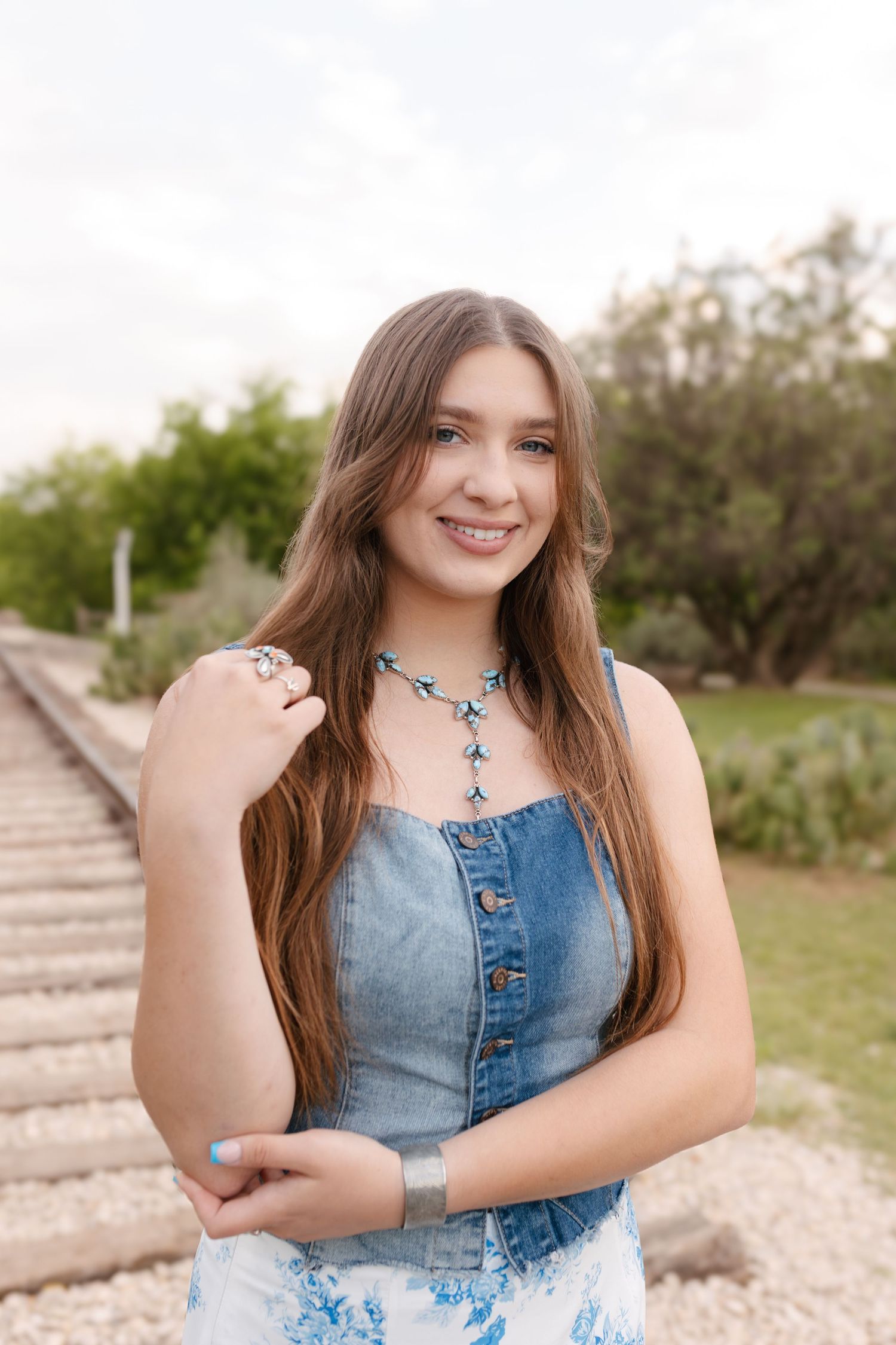 A person in a denim dress poses outdoors on concrete steps with a natural background.