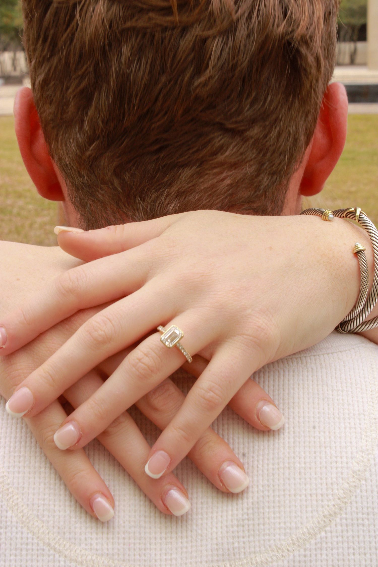 Close-up view of hands wearing an engagement ring resting on someone's shoulder in natural lighting.