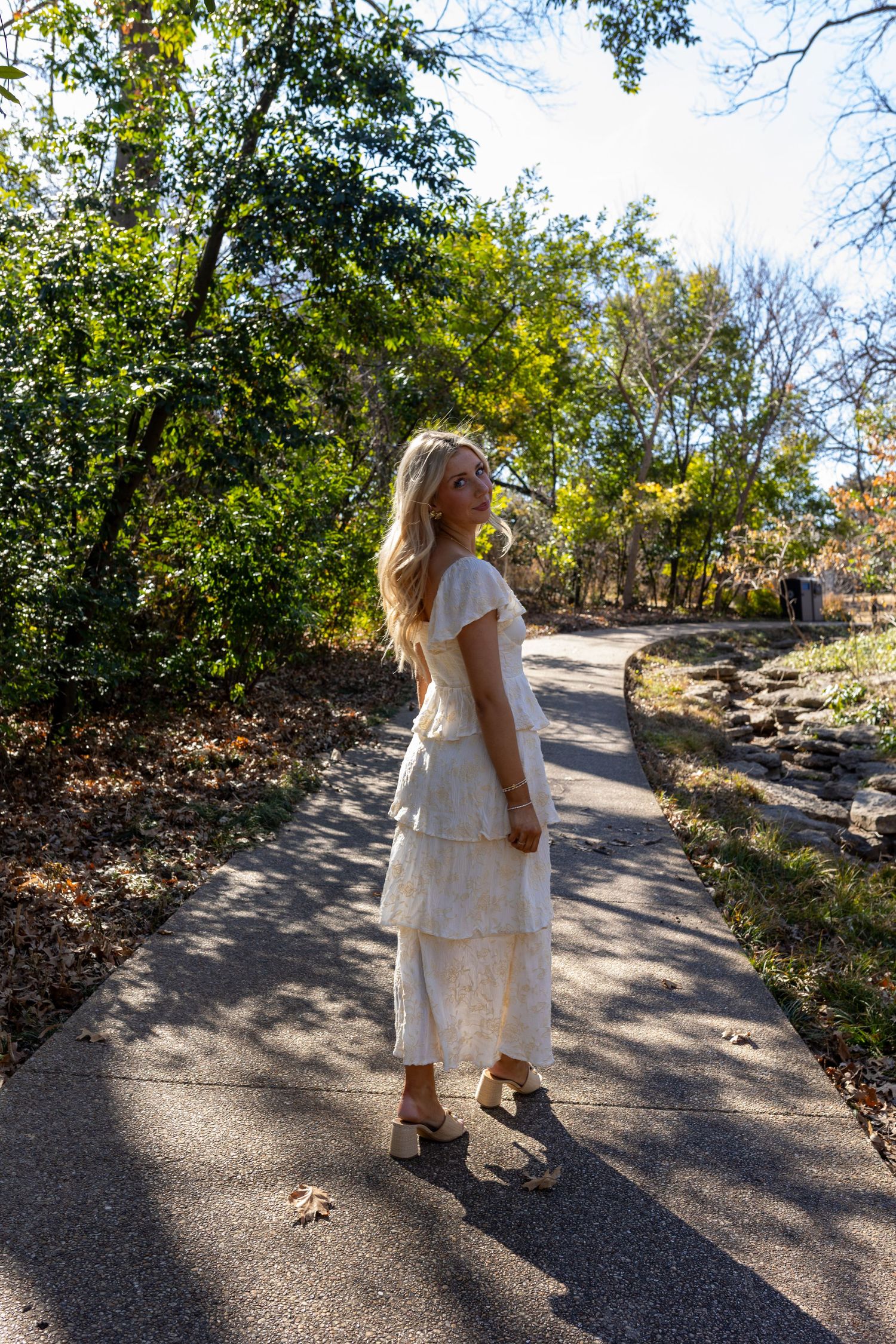 A person in a white dress walks along a winding path through a wooded area on a sunny autumn day.