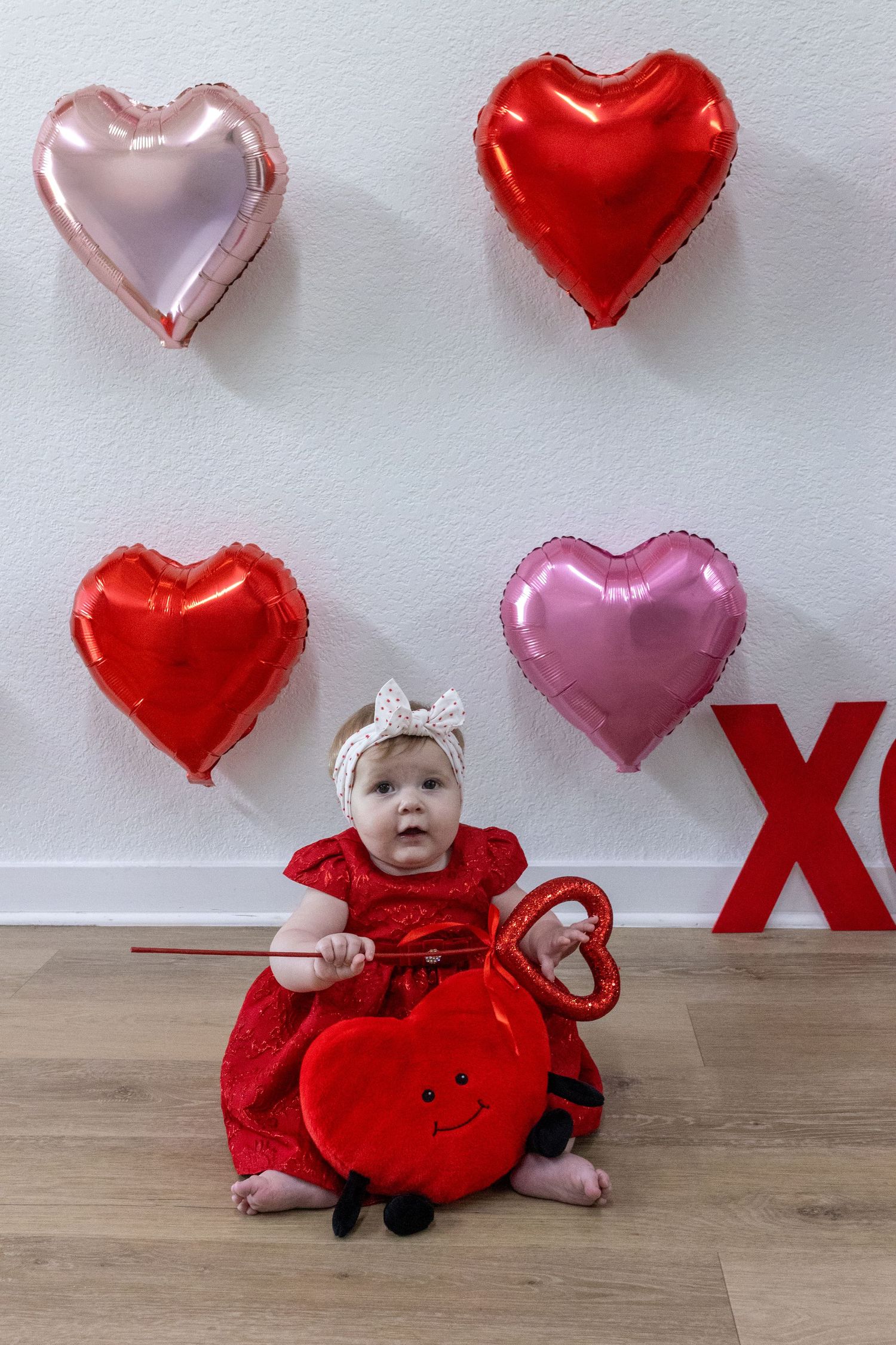 A small figure in a red dress sits on wooden flooring with heart-shaped balloons and decorations in the background.