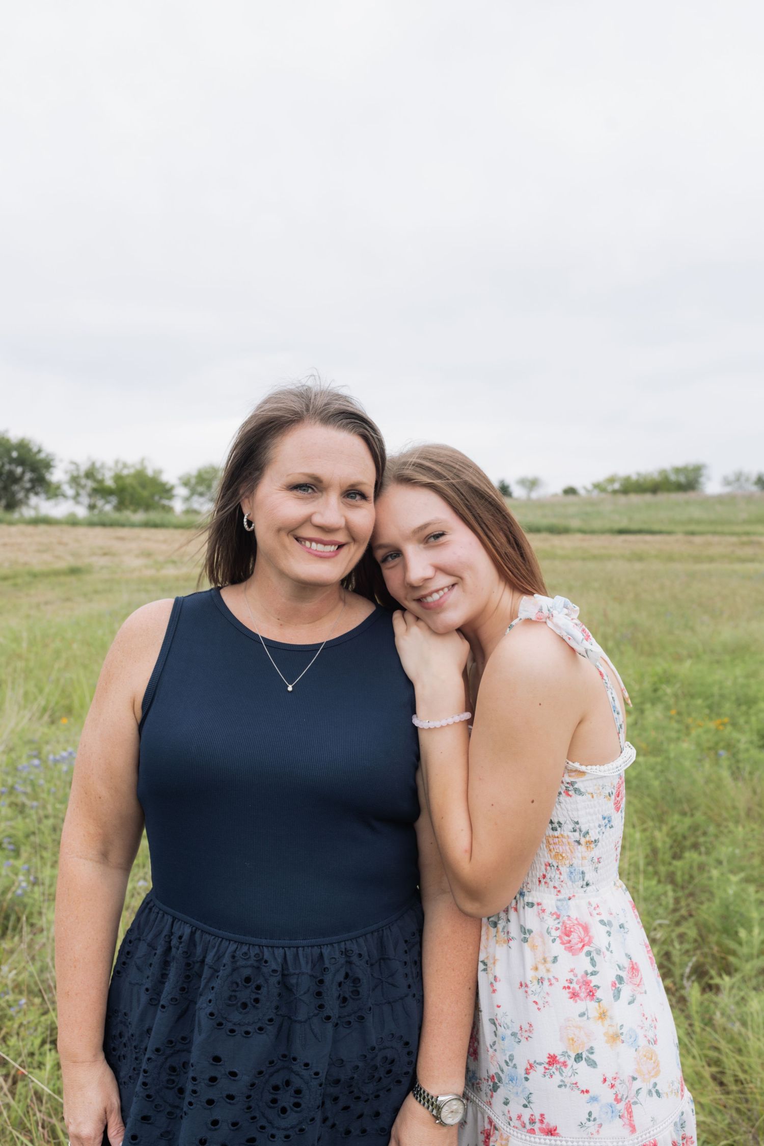 Two women smile together in a green field, one leaning on the others shoulder.