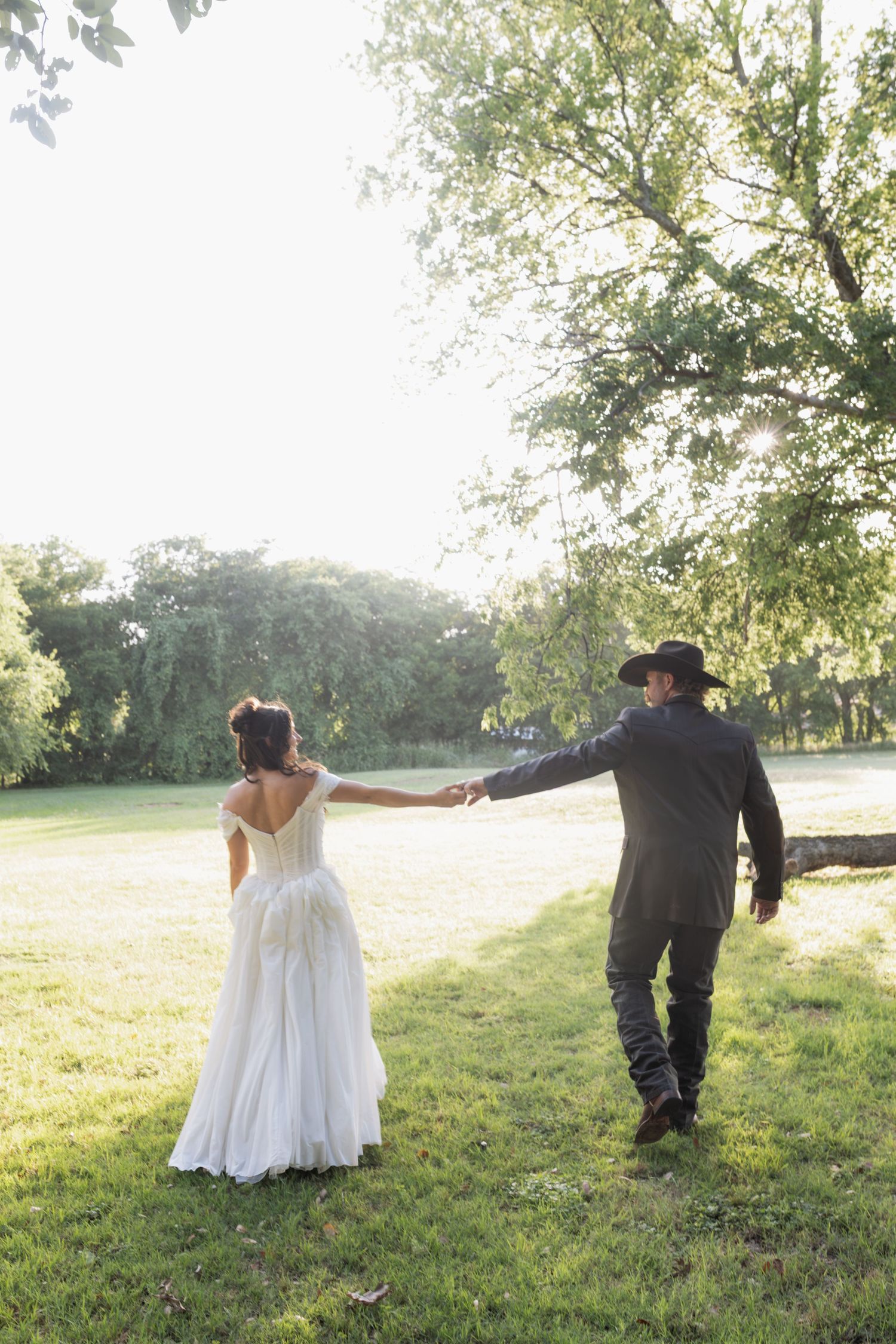 Bride and groom holding hands walking through lush green park on sunny wedding day.