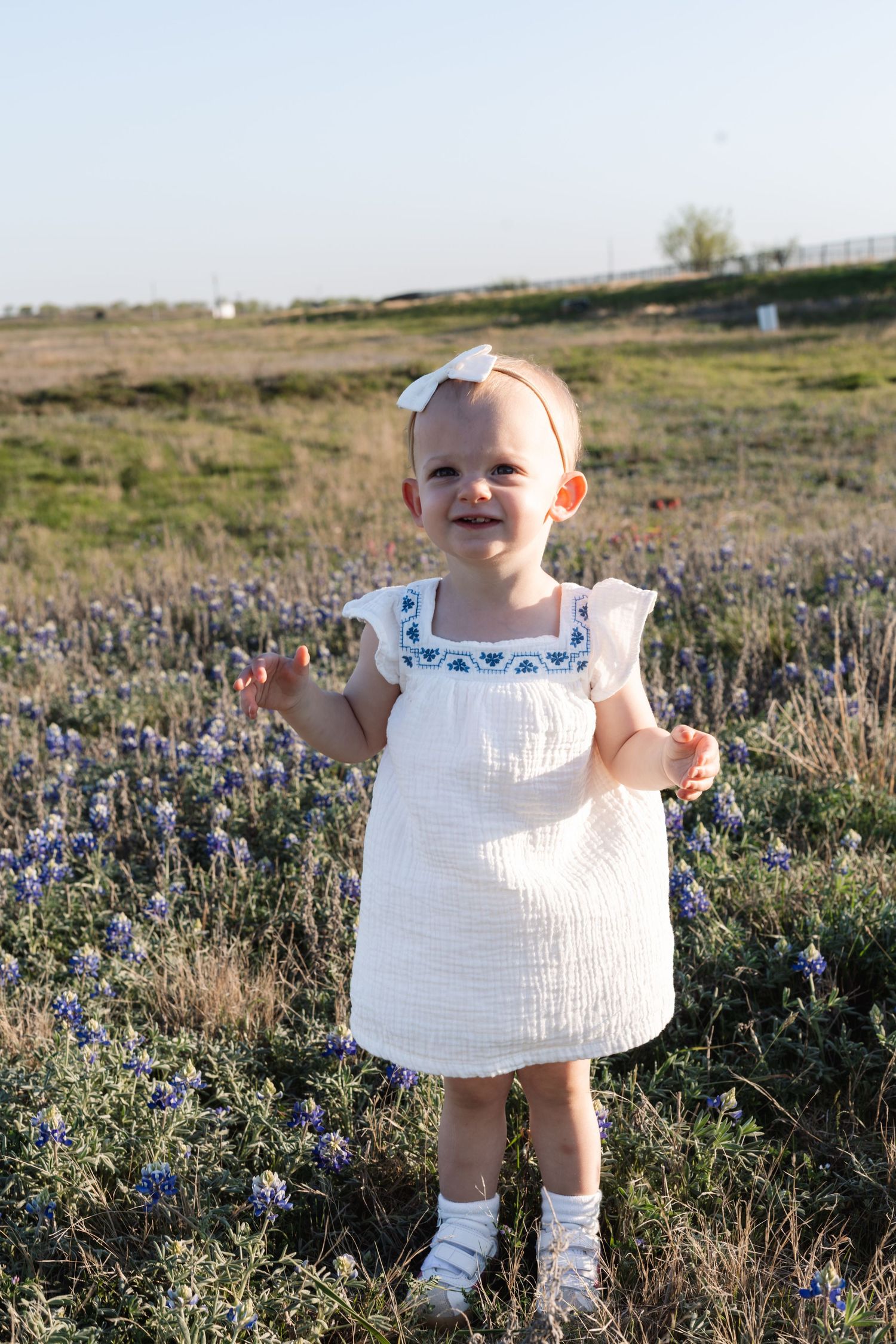 A small child in a white dress stands joyfully among blooming wildflowers in a sunny field.