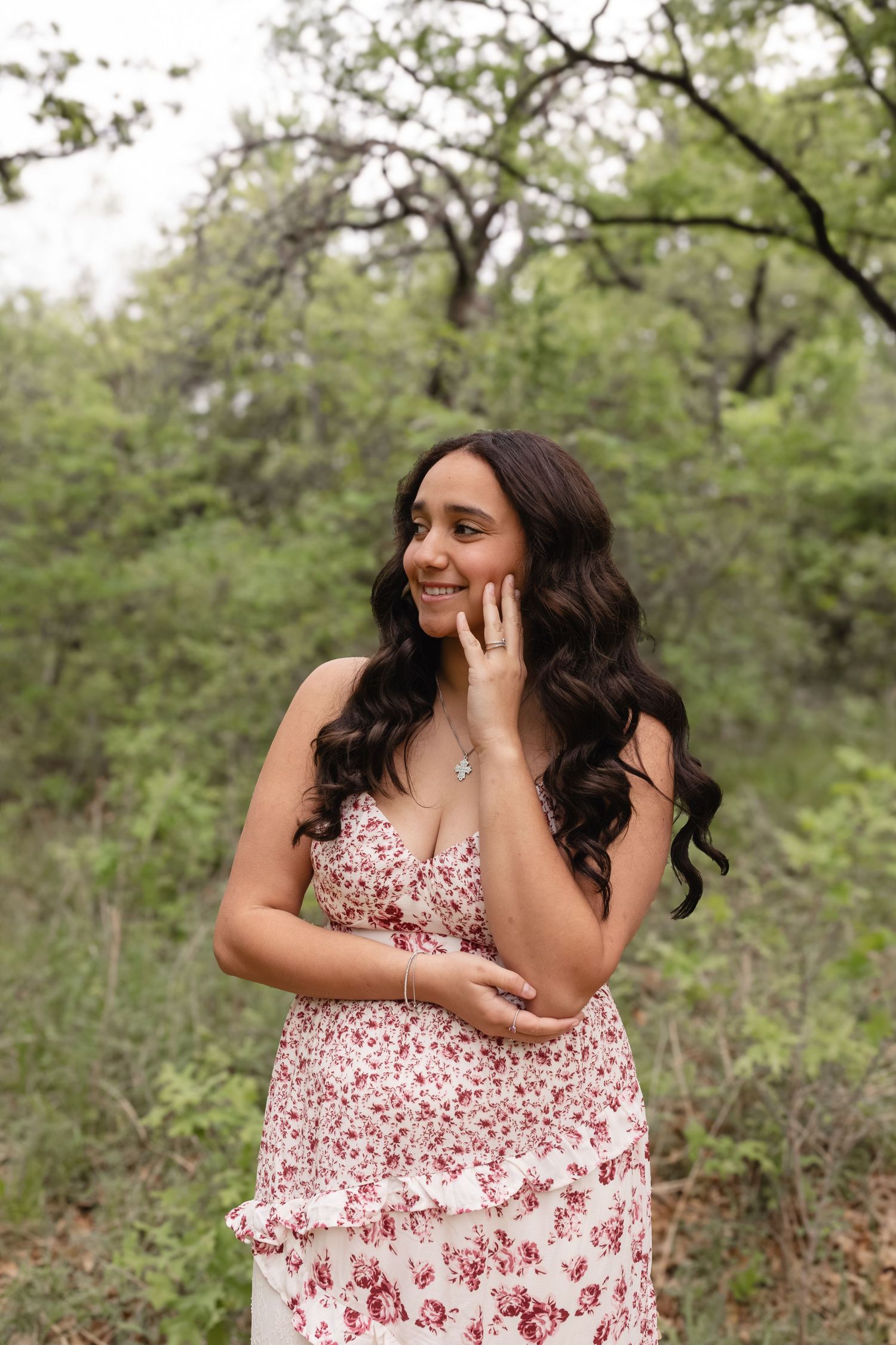 Young woman in floral dress with wavy hair smiling outdoors among lush green trees.