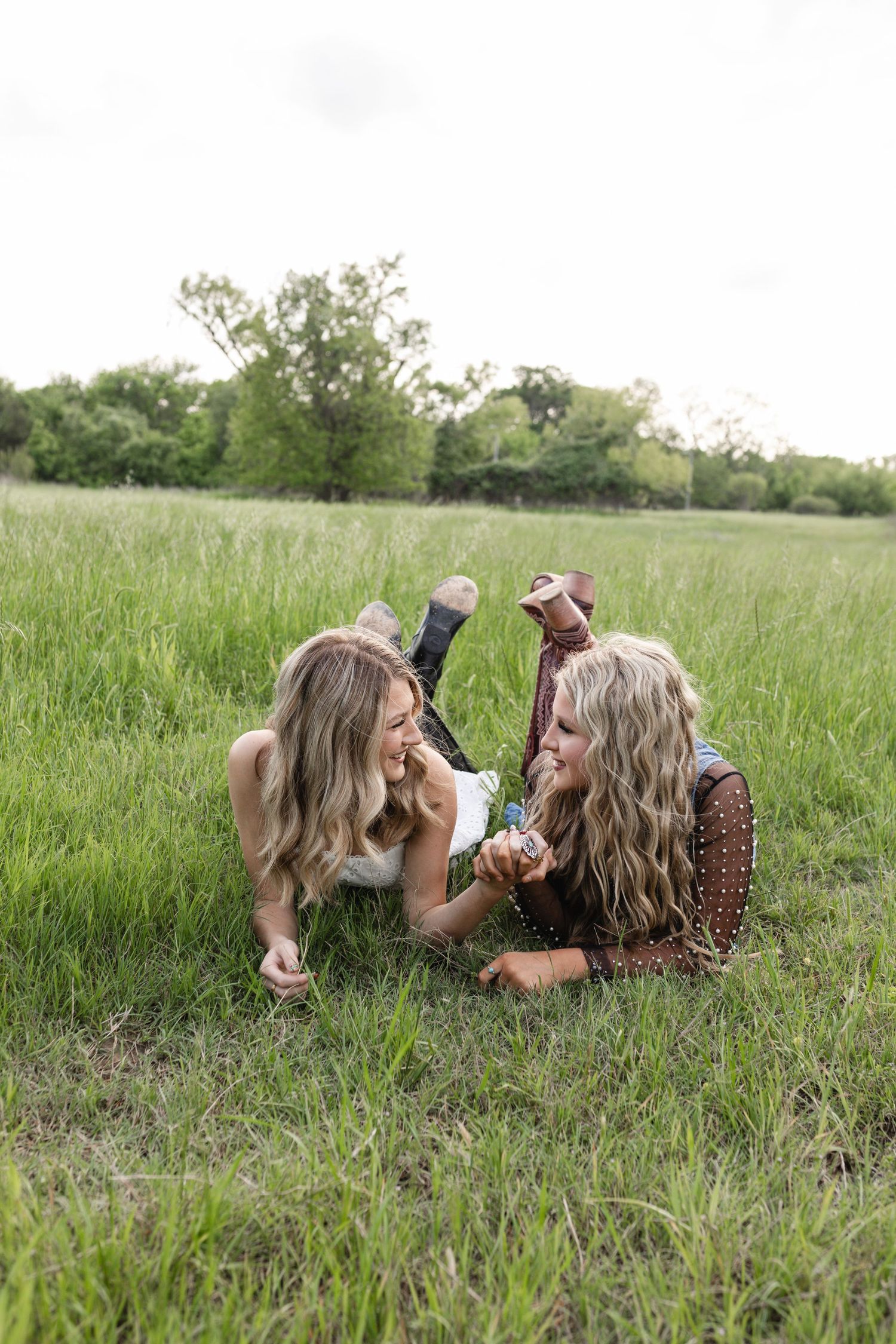 Two blonde women laughing and lying in a lush green field, wearing bohemian outfits and cowboy boots on a cloudy day.
