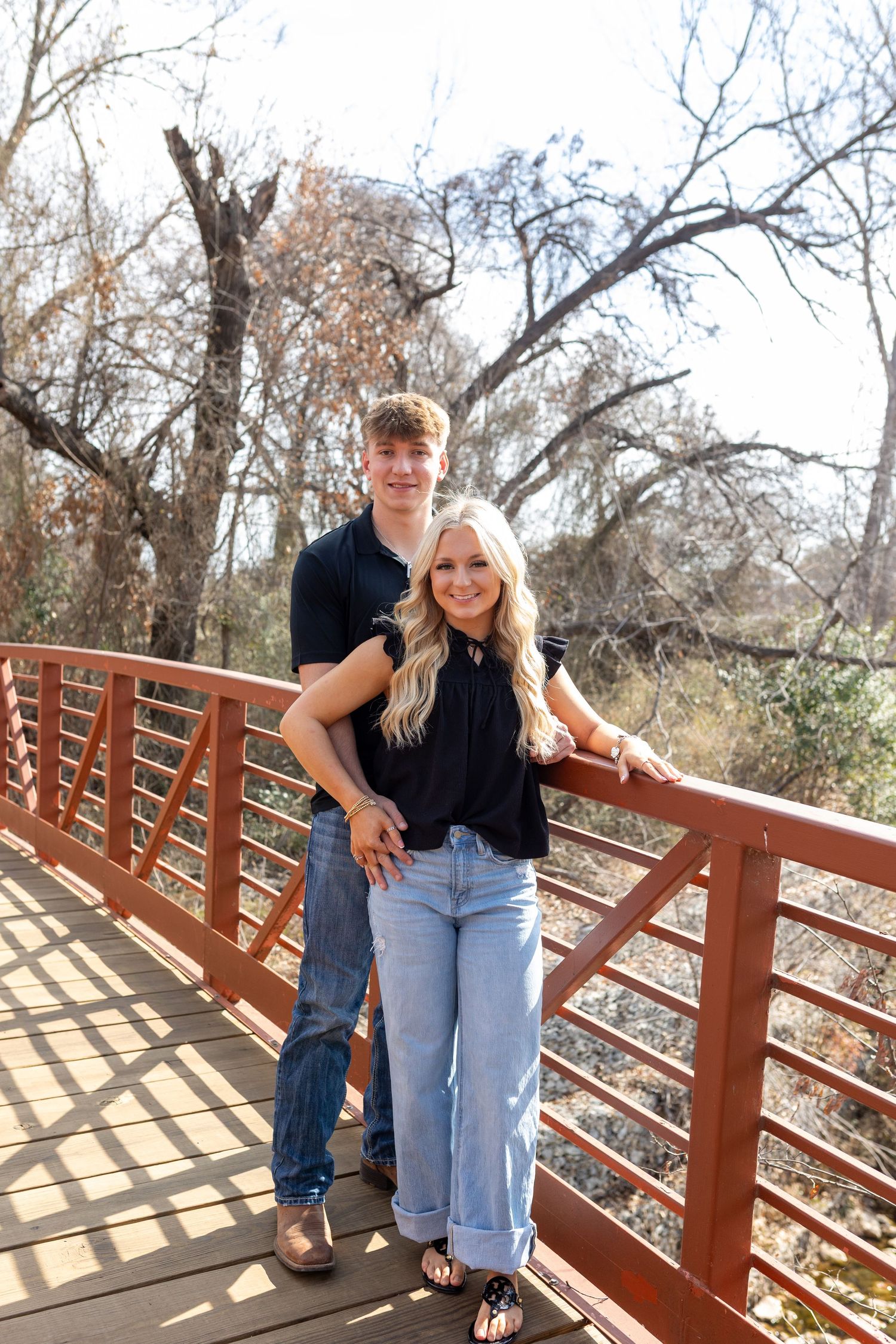 Couple standing together on a wooden bridge walkway surrounded by winter trees.