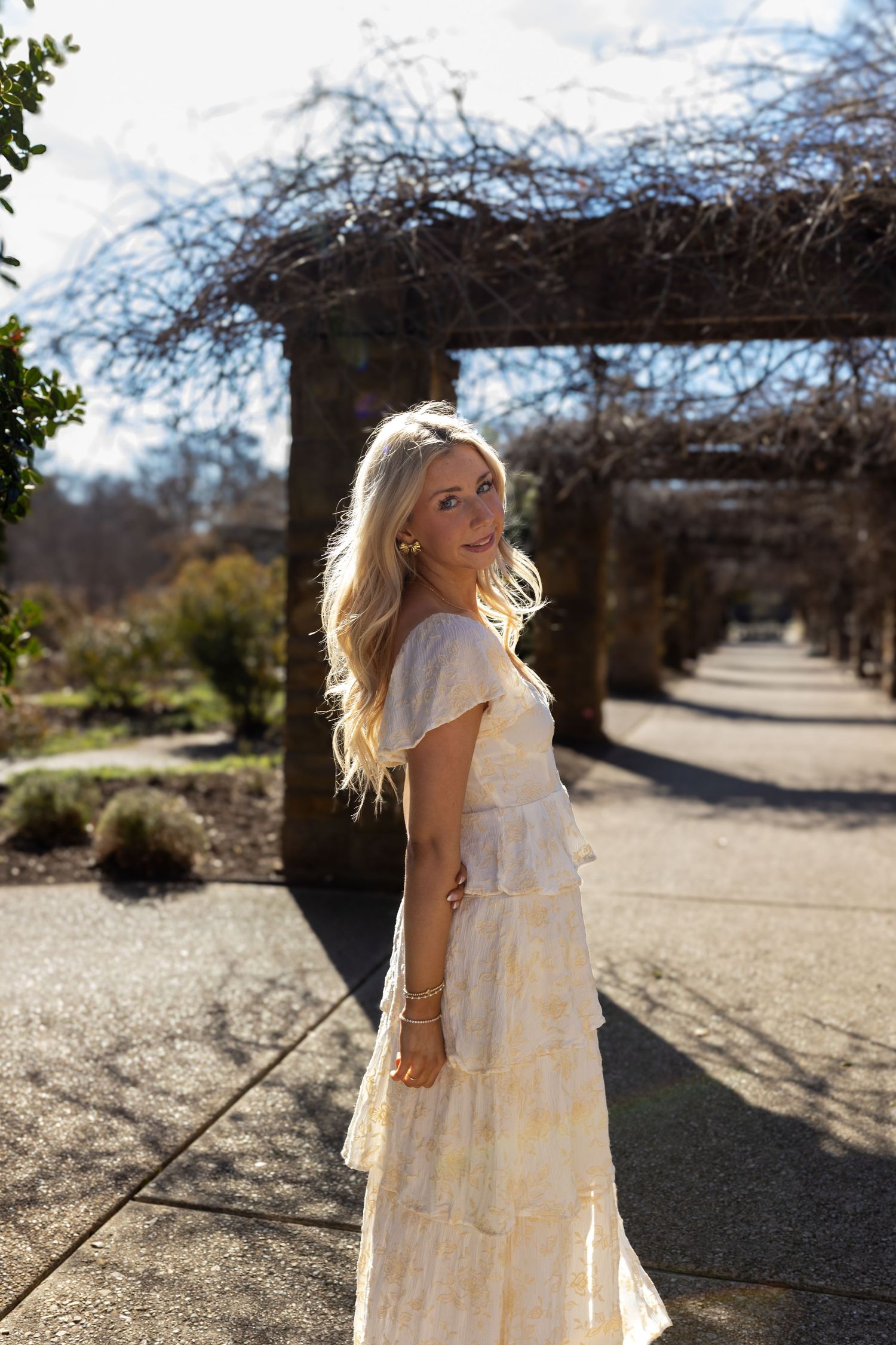 A flowing cream-colored tiered dress is captured in a sunny outdoor garden setting with trees in the background.