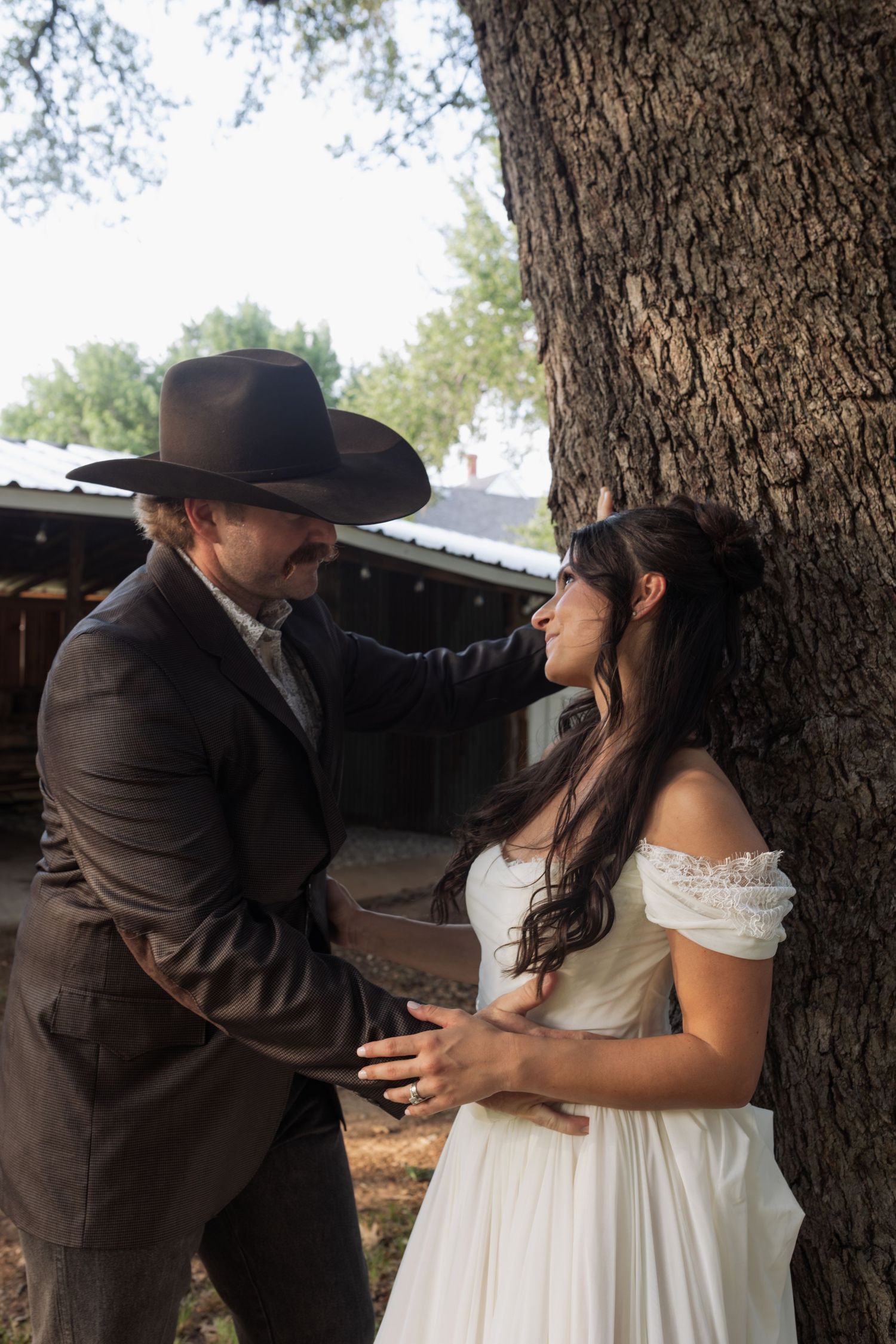 Groom in cowboy hat gazing at bride leaning against tree trunk at rustic outdoor wedding.