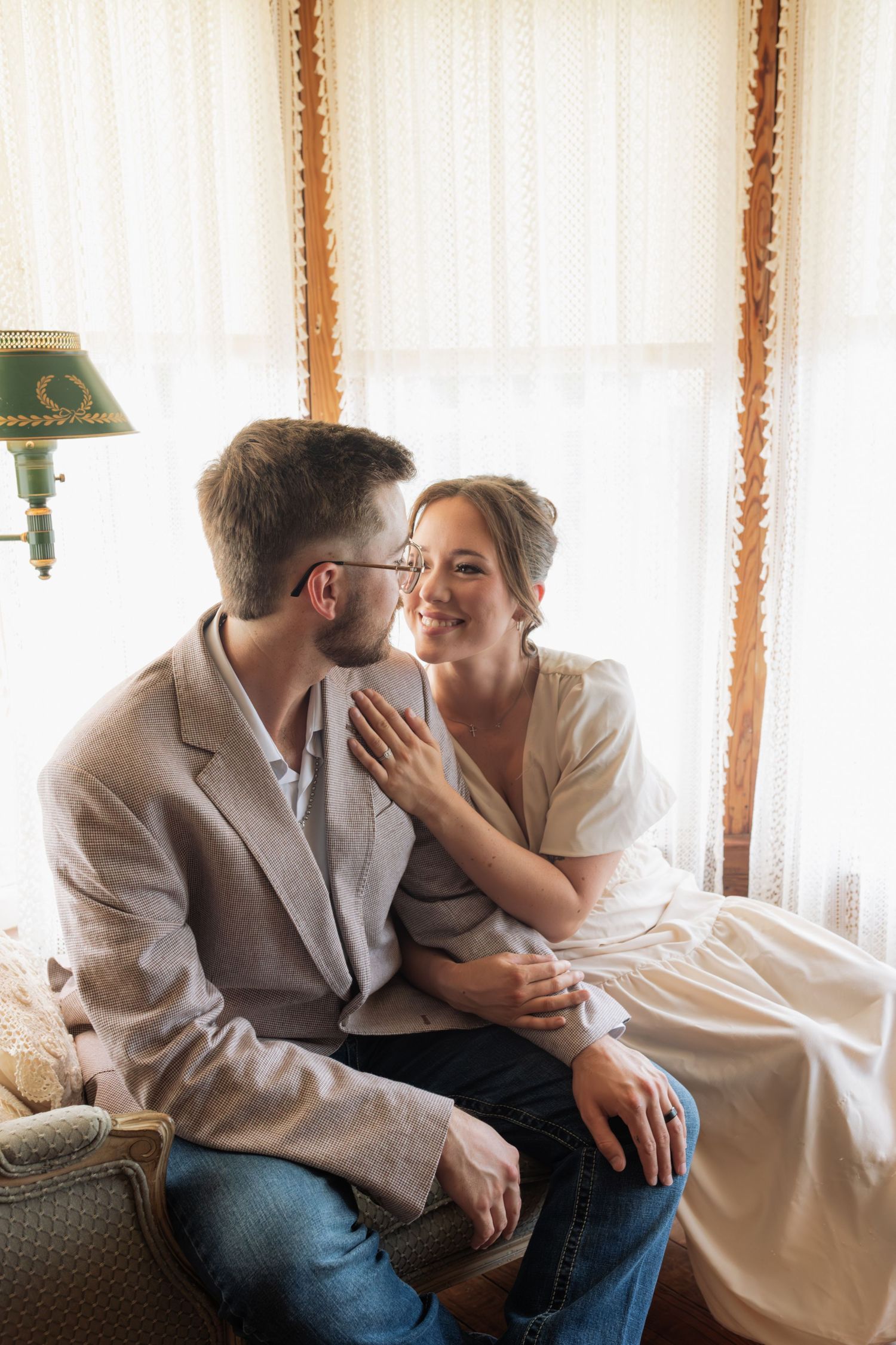A smiling couple sits close together by a sunlit window, the woman resting her hand on the mans chest.