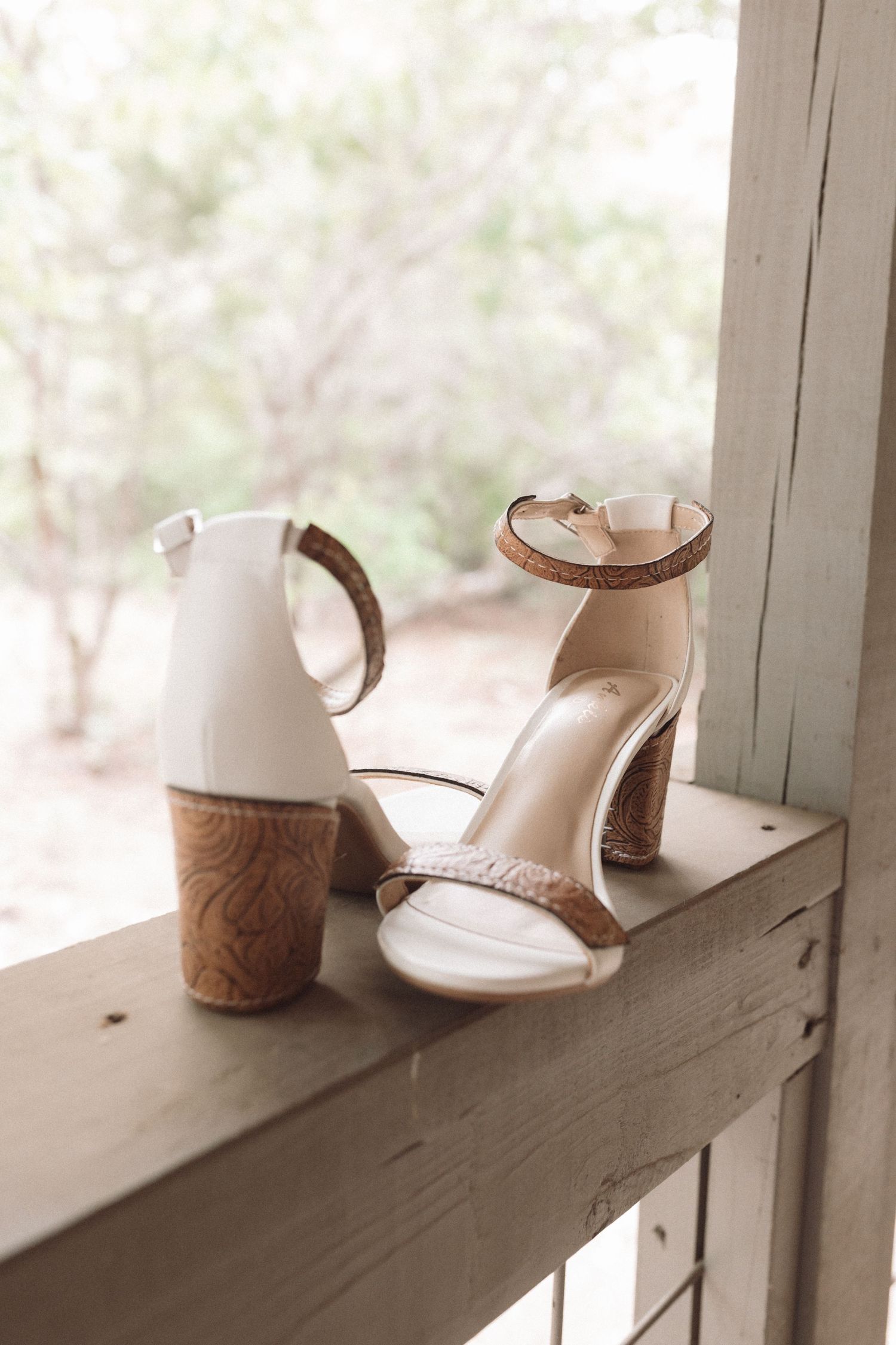 Elegant white and brown cork block heel sandals displayed on a weathered wooden ledge with greenery in the background.