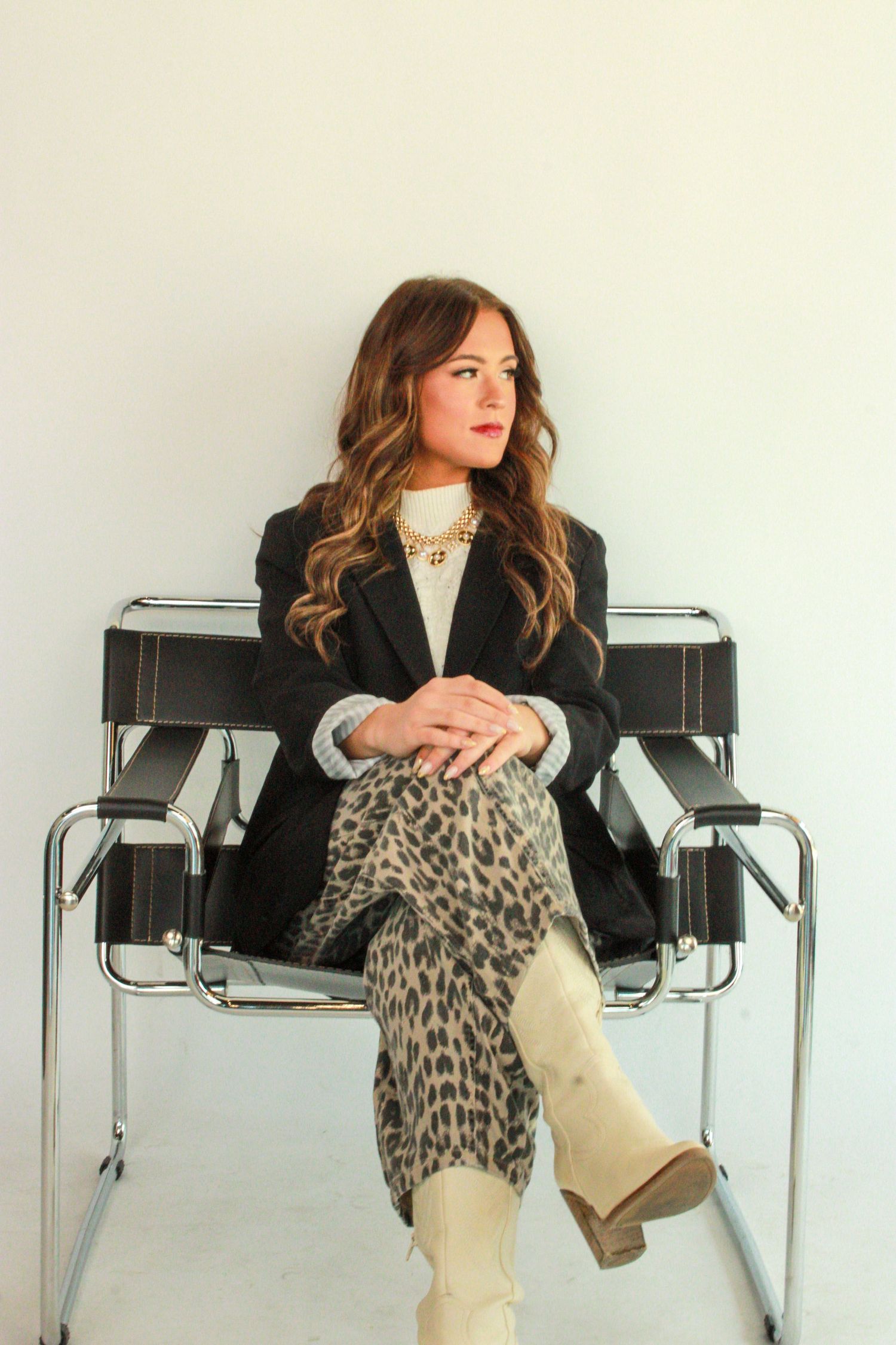 Stylish woman in black blazer and leopard print scarf lounges in modern office chair with cream boots.