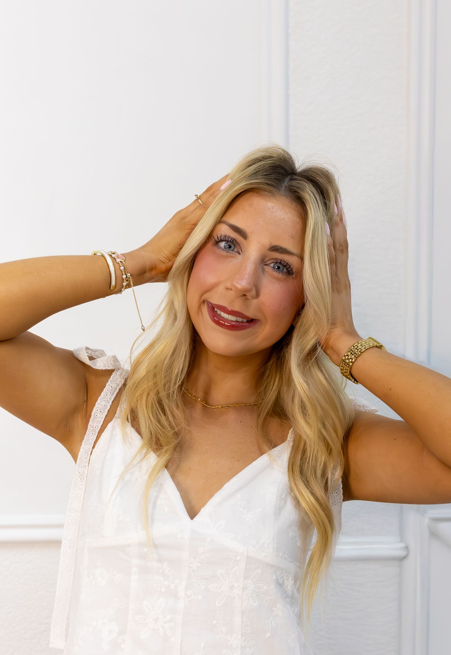 Blonde woman in white dress poses with hands in hair, wearing gold jewelry and bracelets against white wall.