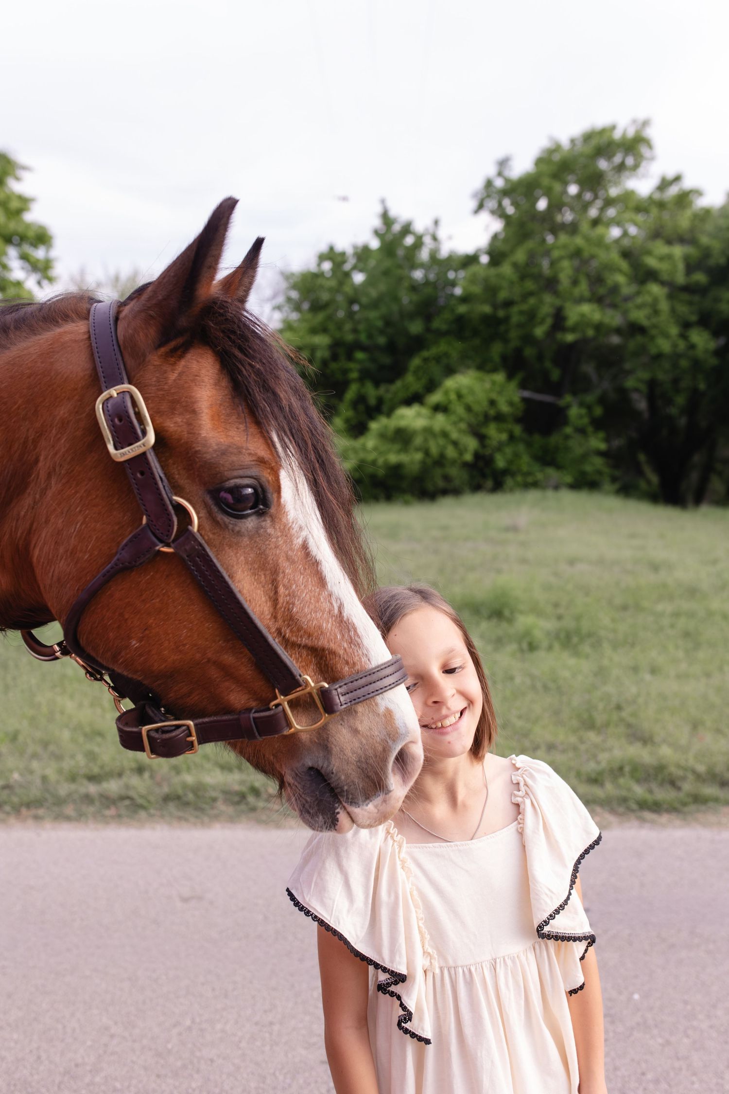 A smiling girl in a white dress stands beside a brown horse with a white blaze wearing a leather halter outdoors.