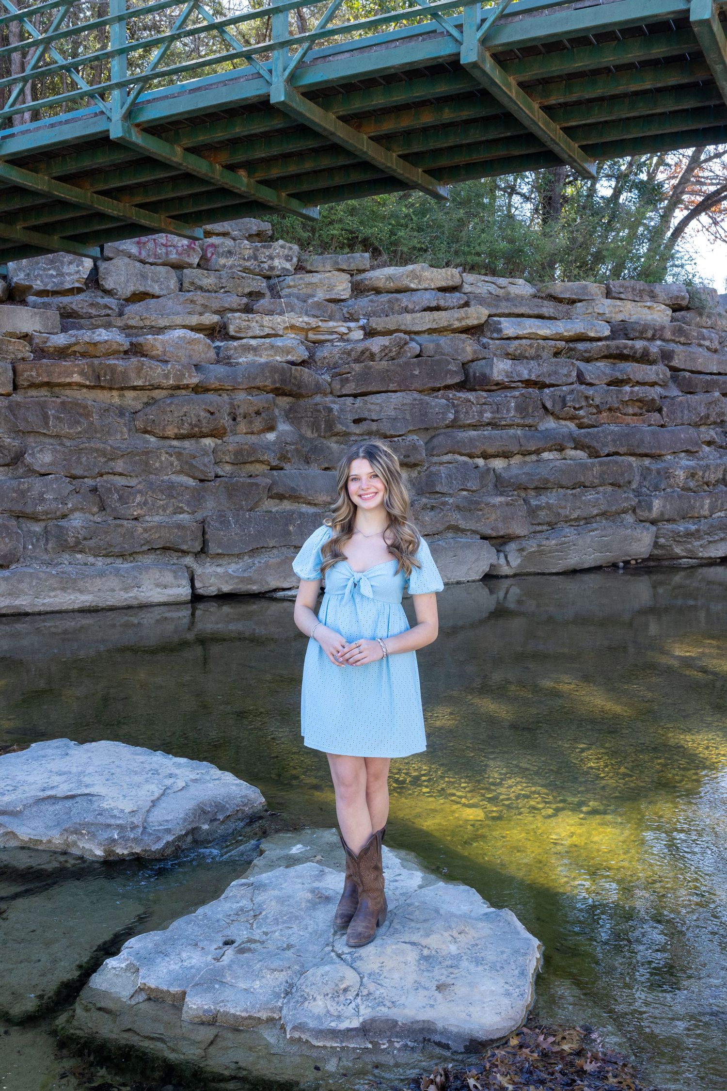 A person in a light blue dress and brown boots poses on rocks near a creek with a stone bridge in the background.