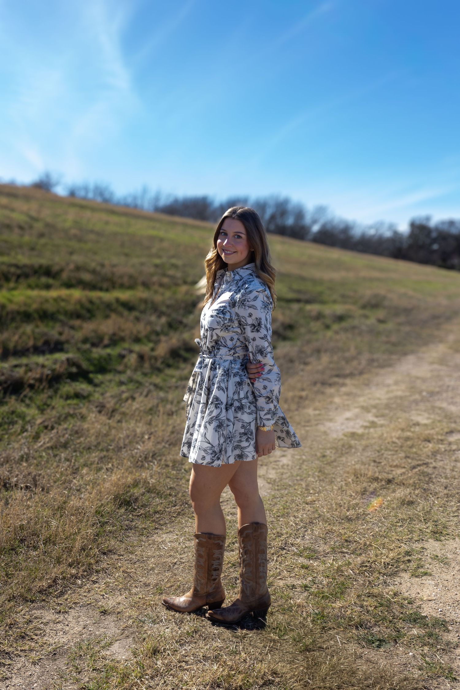 A person in a floral dress and brown boots stands on a dirt path against a blue sky and grassy hillside.