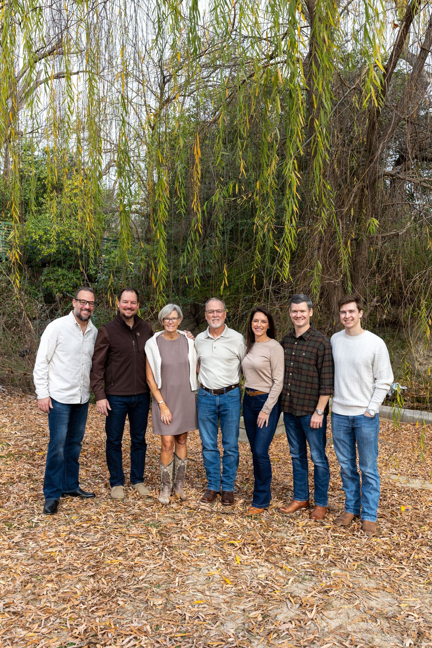 A group of people in casual clothing stand together against a backdrop of willow trees and fallen autumn leaves.