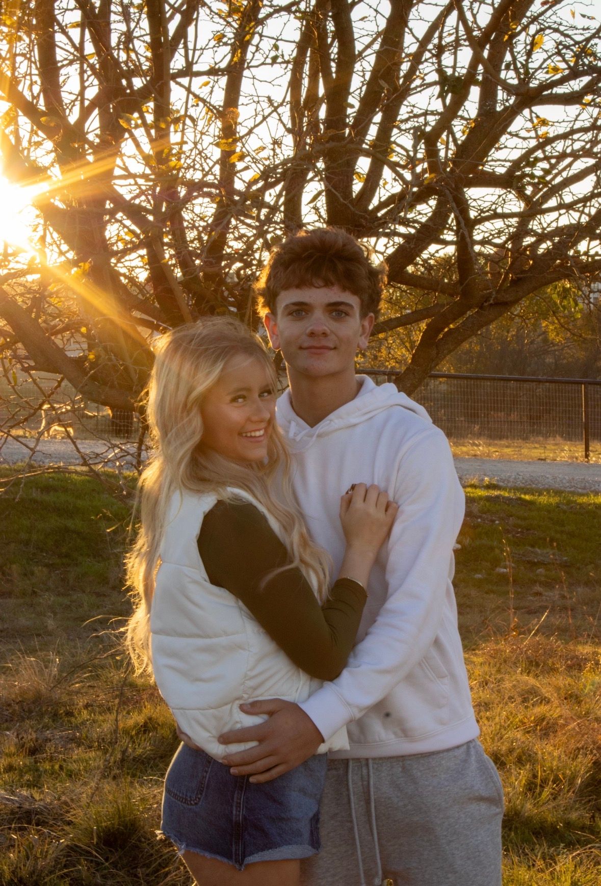 Couple embraces in warm golden sunlight with bare trees and grass in background during sunset.